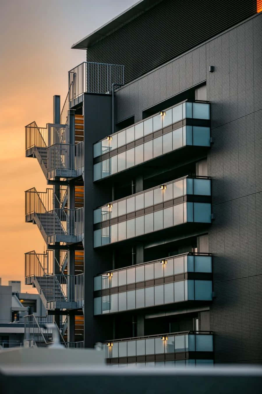 Architecture 桌布 壁紙 — 現代公寓大樓，設有玻璃陽台和金屬防火梯，映襯溫暖的日落天空。