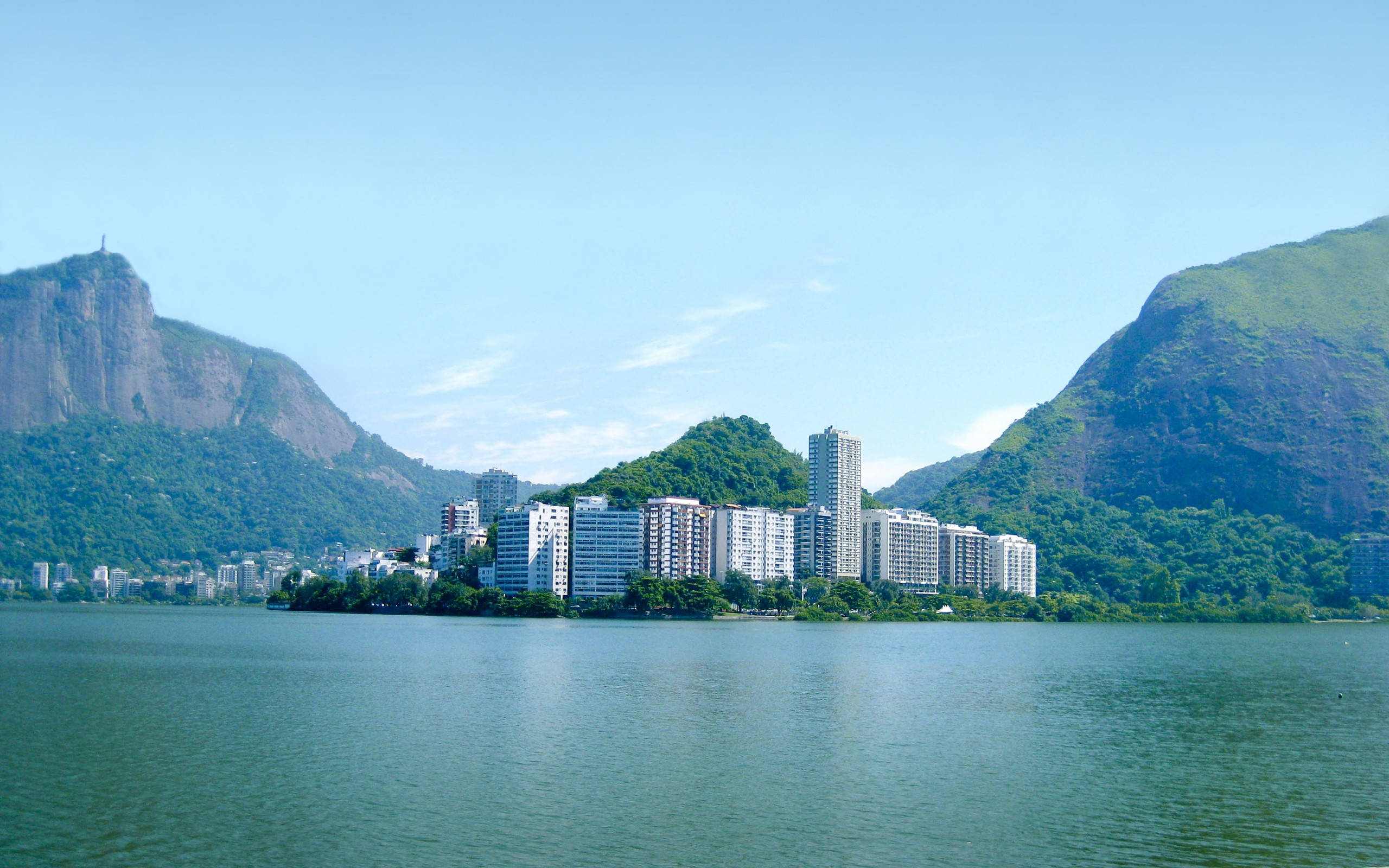 rio 桌布 壁紙 — 現代公寓大樓沿著寧靜的綠色湖泊排列，兩側是茂密的山脈，上方是清澈的藍天。