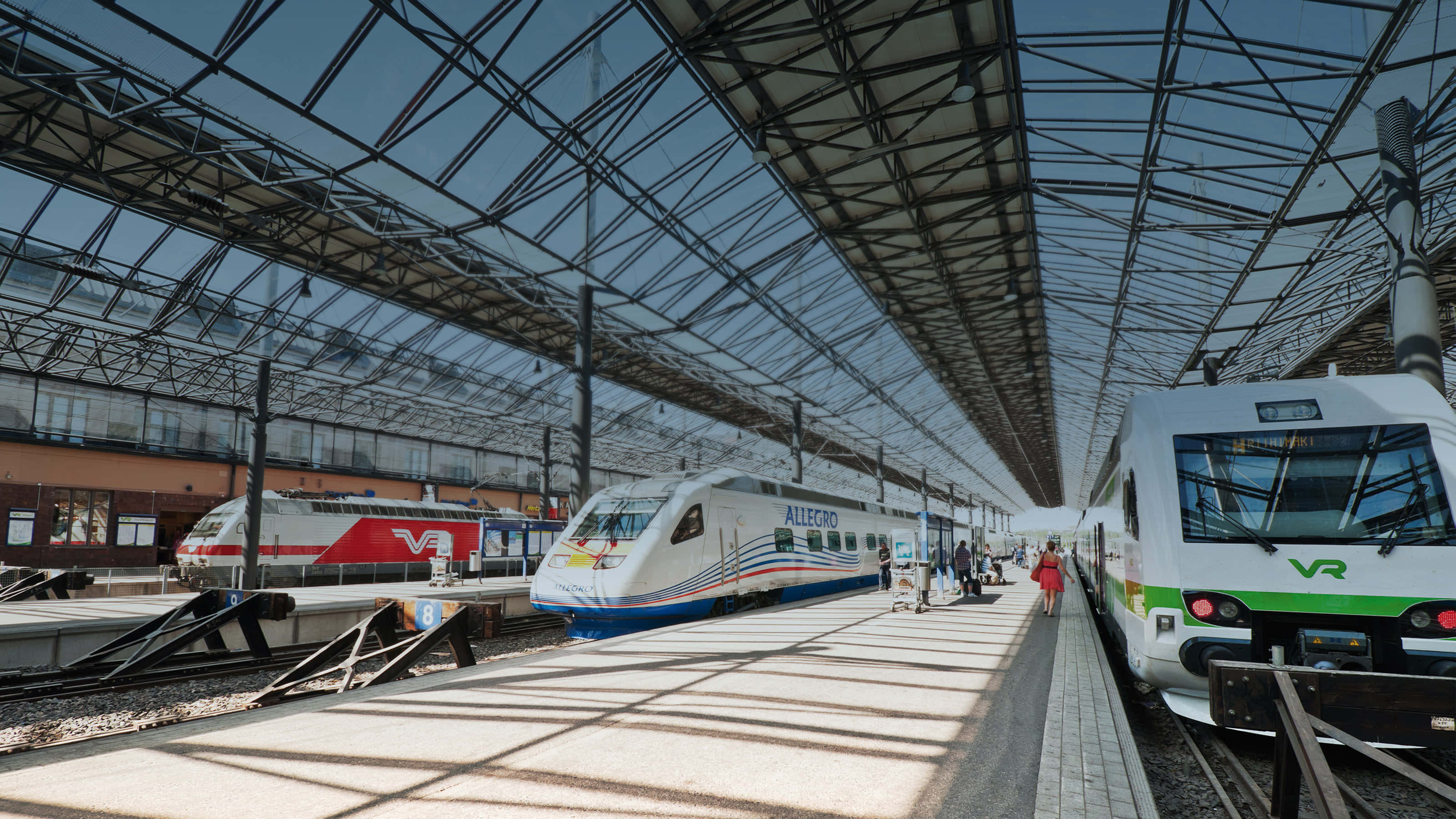 Train Station 桌布 壁紙 — 現代列車停泊在寬闊的玻璃和鋼結構車站屋頂下的月台上，陽光投下長長的影子。