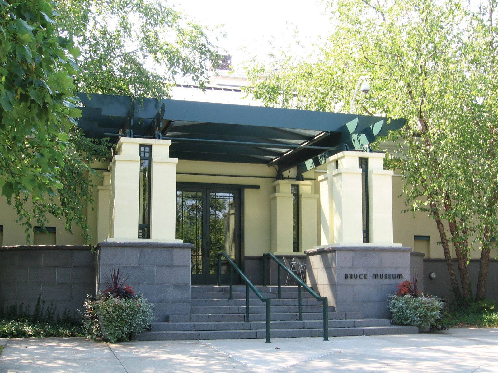 Museum 桌布 壁紙 — 現代博物館入口，有奶油色柱子、綠色欄杆和石階，周圍環繞著茂密的綠樹，在明亮的日光下。