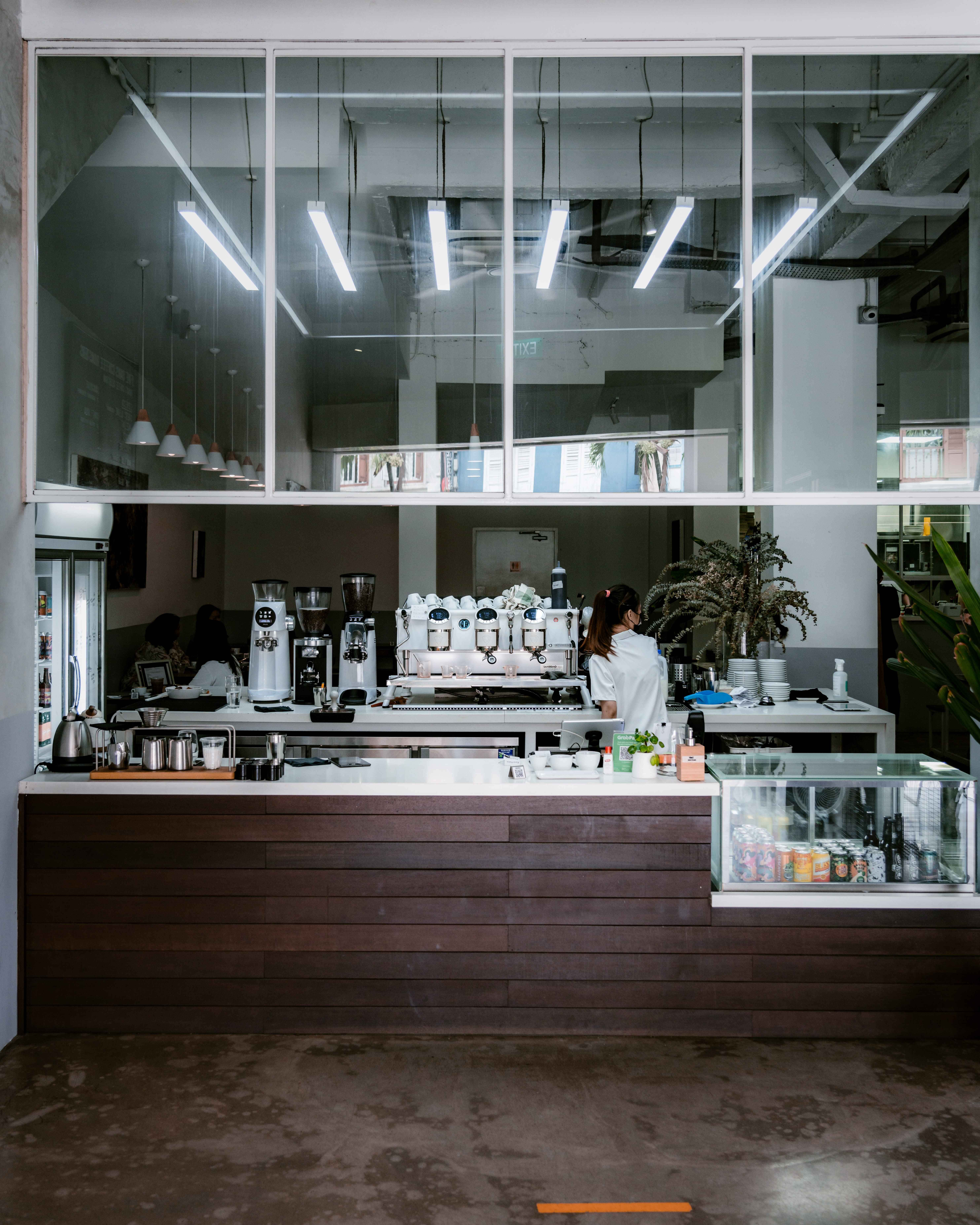 Barista 桌布 壁紙 — 現代咖啡店內部，咖啡師在明亮天花燈下於時尚木製櫃檯後工作。