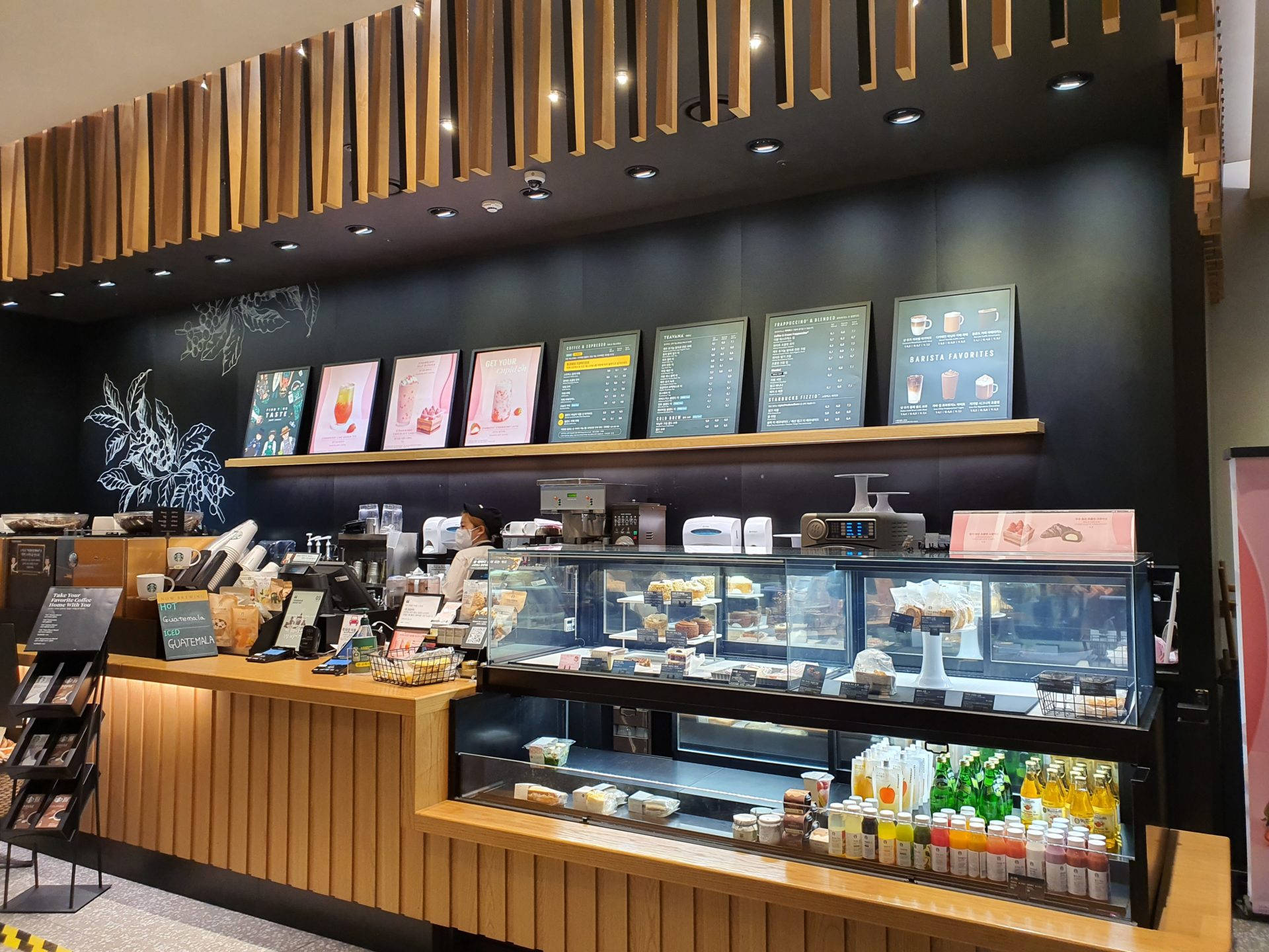 咖啡動圖 桌布 壁紙 — 現代咖啡店內部，木質裝飾、黑色牆壁、菜單板，以及裝滿糕點和瓶裝飲料的玻璃展示櫃。