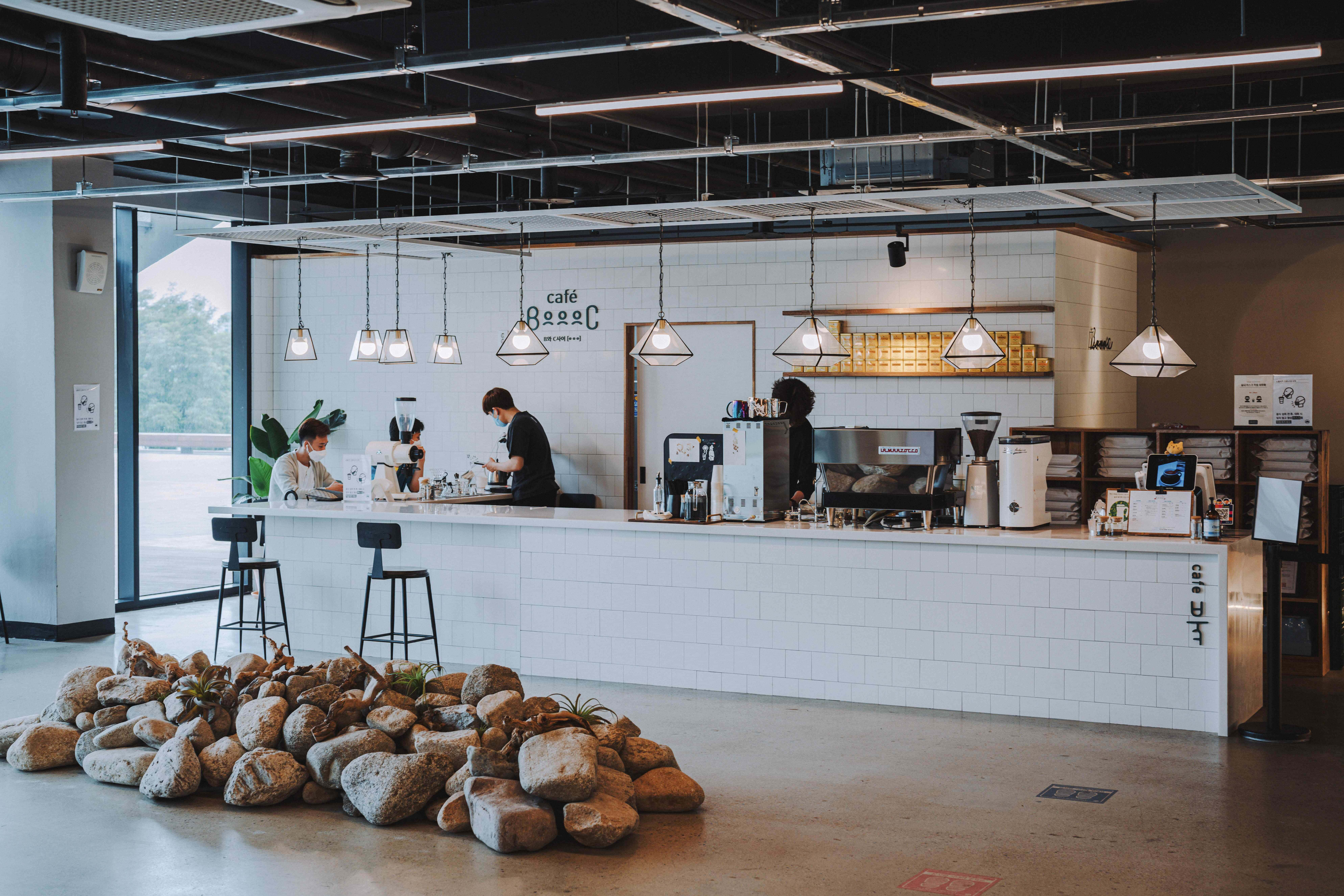 Coffee Aesthetic 桌布 壁紙 — 現代咖啡館內部，設有白色瓷磚吧檯，員工和顧客在工業吊燈下，前景有大型岩石佈置。