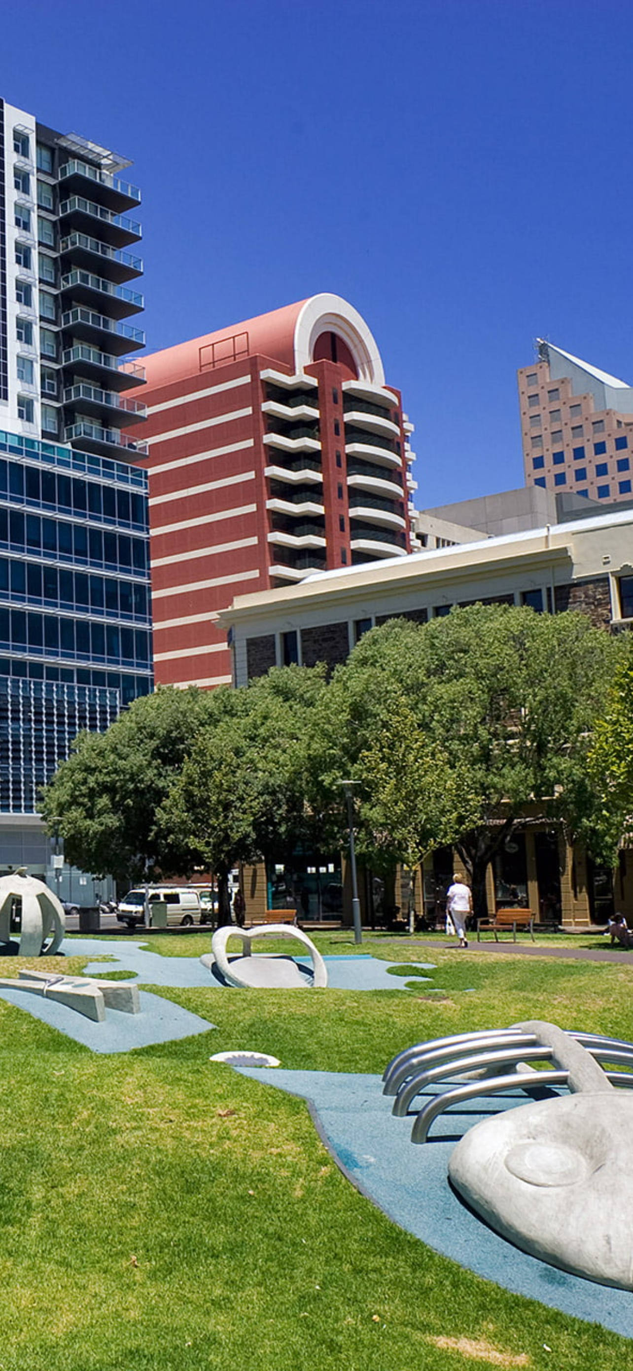 Adelaide 桌布 壁紙 — 現代城市公園，在晴朗的藍天下，周圍是高樓大廈和茂密的綠樹，設有雕塑般的遊樂場設備。