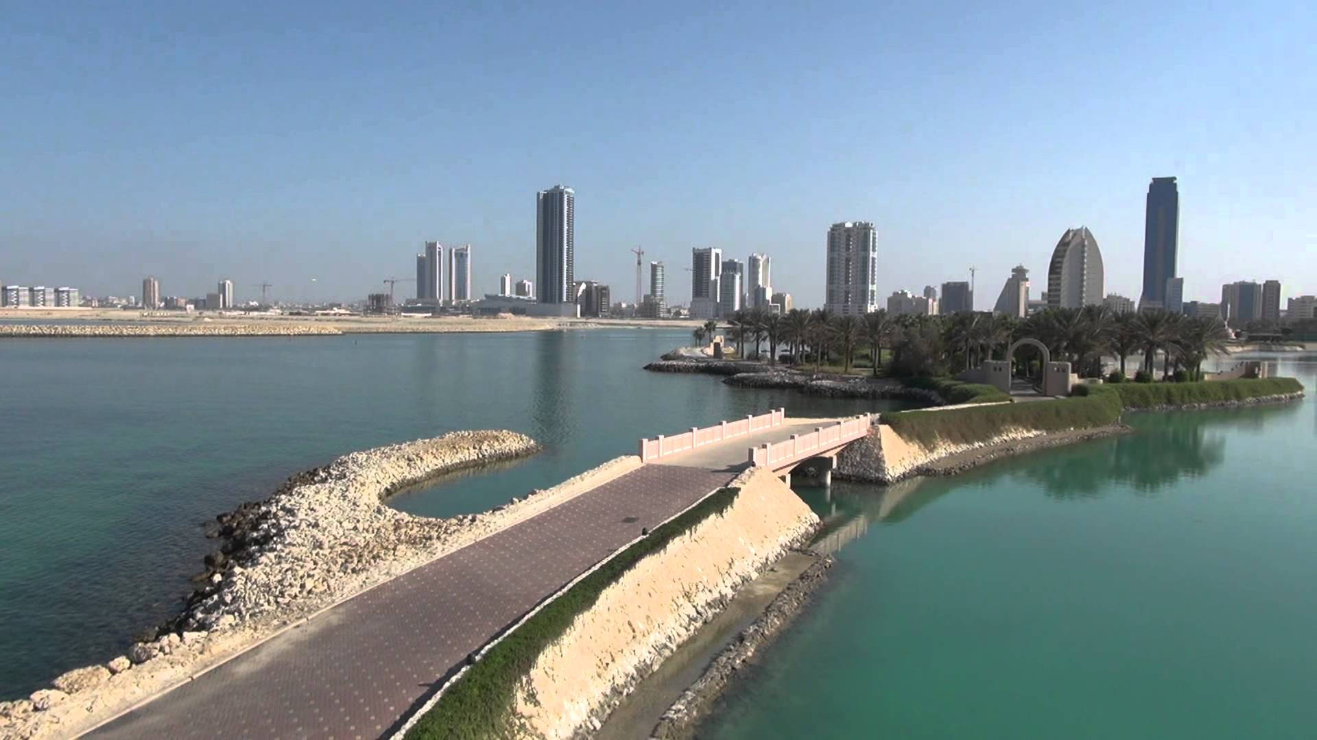 Bahrain 桌布 壁紙 — 現代城市天際線在碧綠水道後方升起，水道上有彎曲橋樑和棕櫚樹成蔭的步道，天空清澈湛藍。