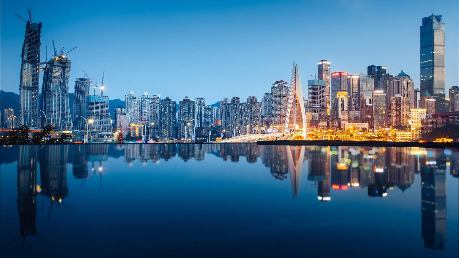 Chongqing 桌布 壁紙 — 現代城市天際線在黃昏時分，燈火通明的摩天大樓和橋樑倒映在平靜的水面上。