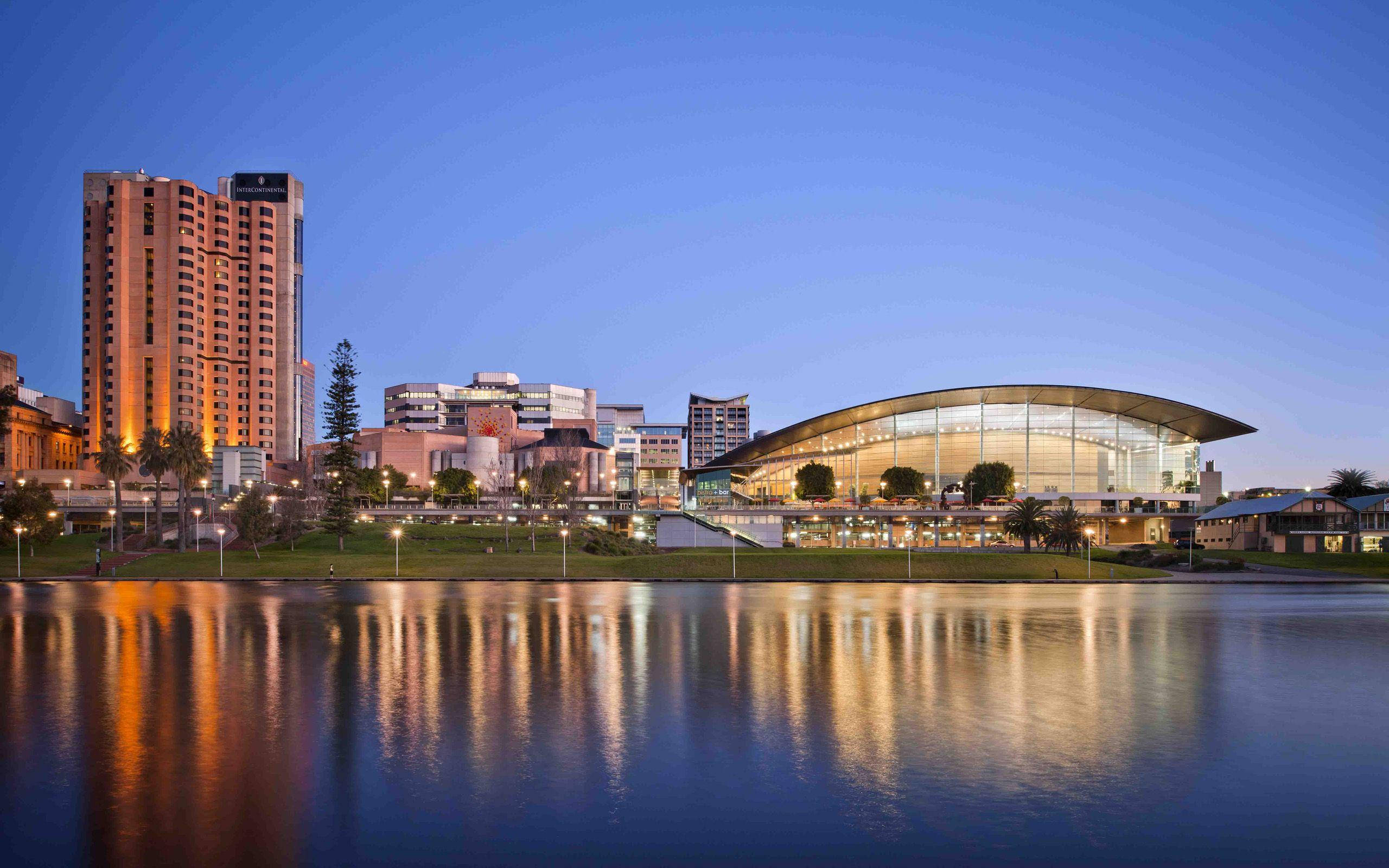 Adelaide 桌布 壁紙 — 現代城市天際線在黃昏時分，燈火通明的建築物倒映在平靜的水面上。