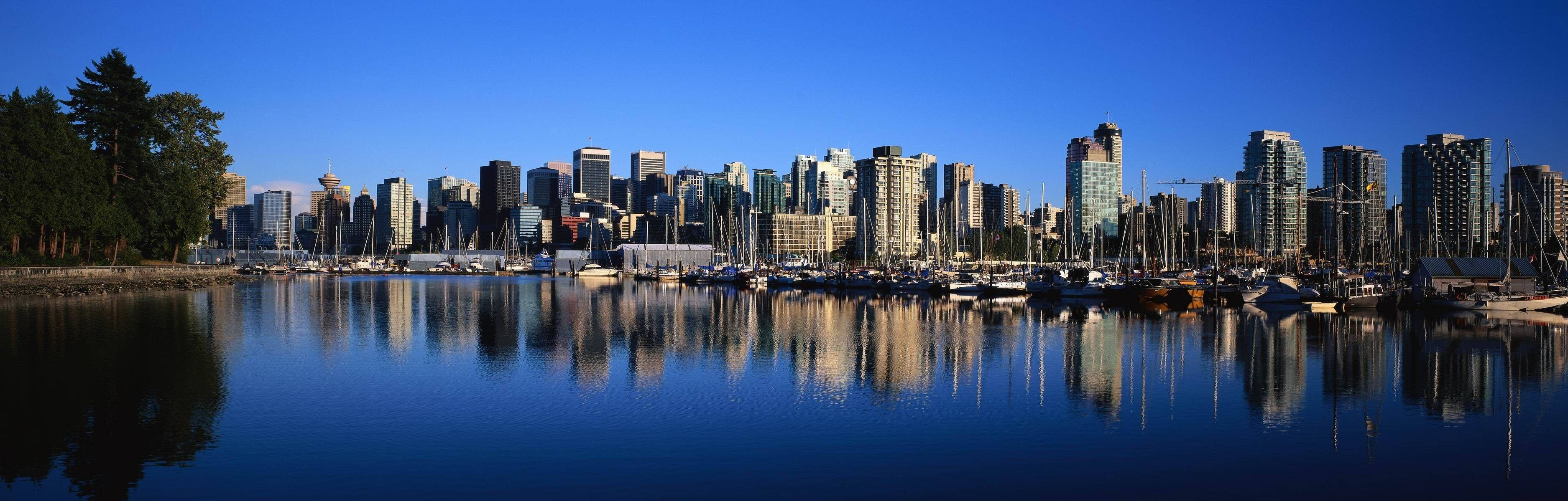 Blue Sky 桌布 壁紙 — 現代城市天際線的全景視圖，完美倒映在平靜的藍色水面上，前景有停泊的帆船。