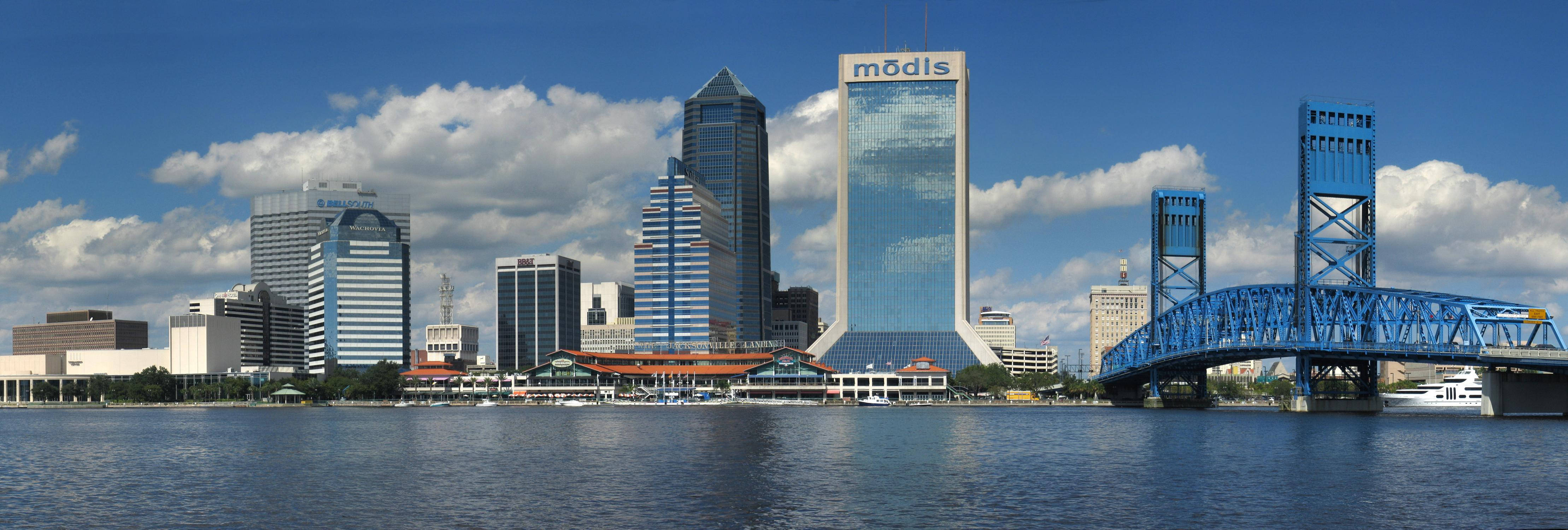 Jacksonville 桌布 壁紙 — 現代城市天際線的全景視圖，玻璃摩天大樓和藍色橋樑橫跨平靜的水面，天空部分多雲。