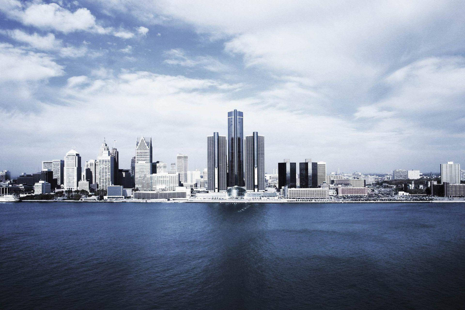 detroit 桌布 壁紙 — 現代城市天際線的全景視圖，高聳的摩天大樓在部分多雲的天空下倒映在平靜的藍色水面上。