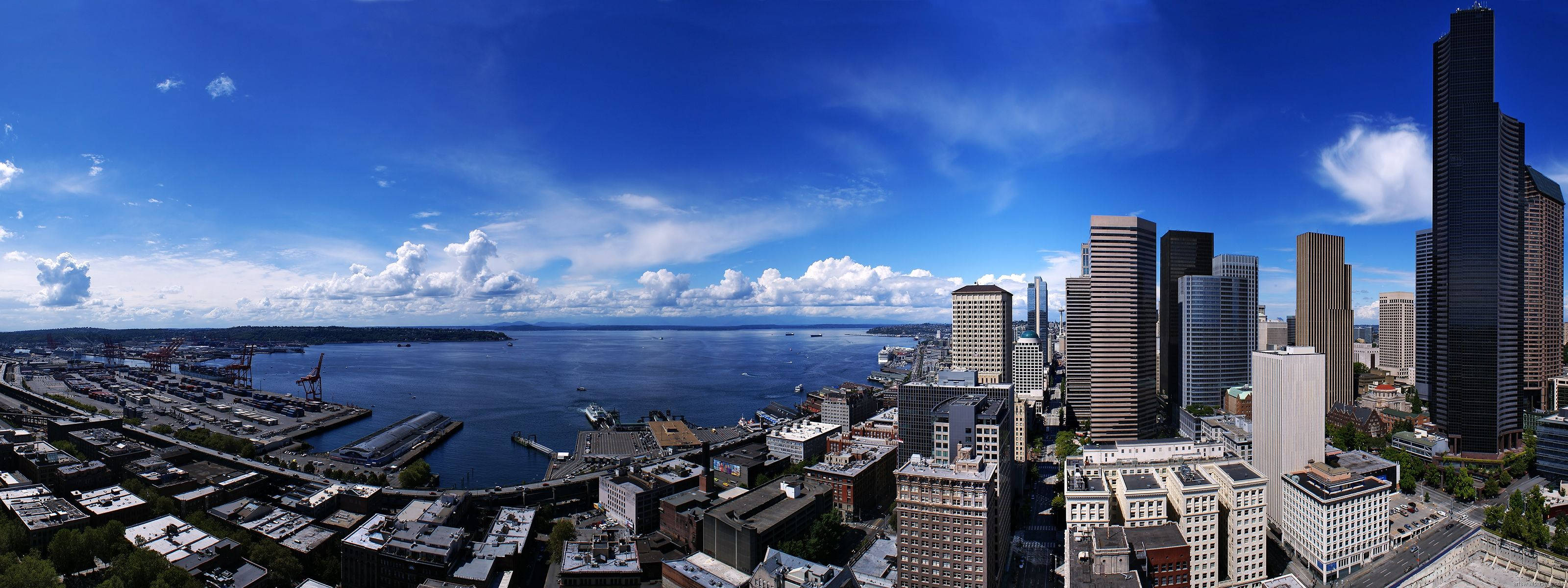Seattle Skyline 桌布 壁紙 — 現代城市天際線的全景視圖，高聳的摩天大樓和港口，在明亮的藍天和蓬鬆的白雲下。