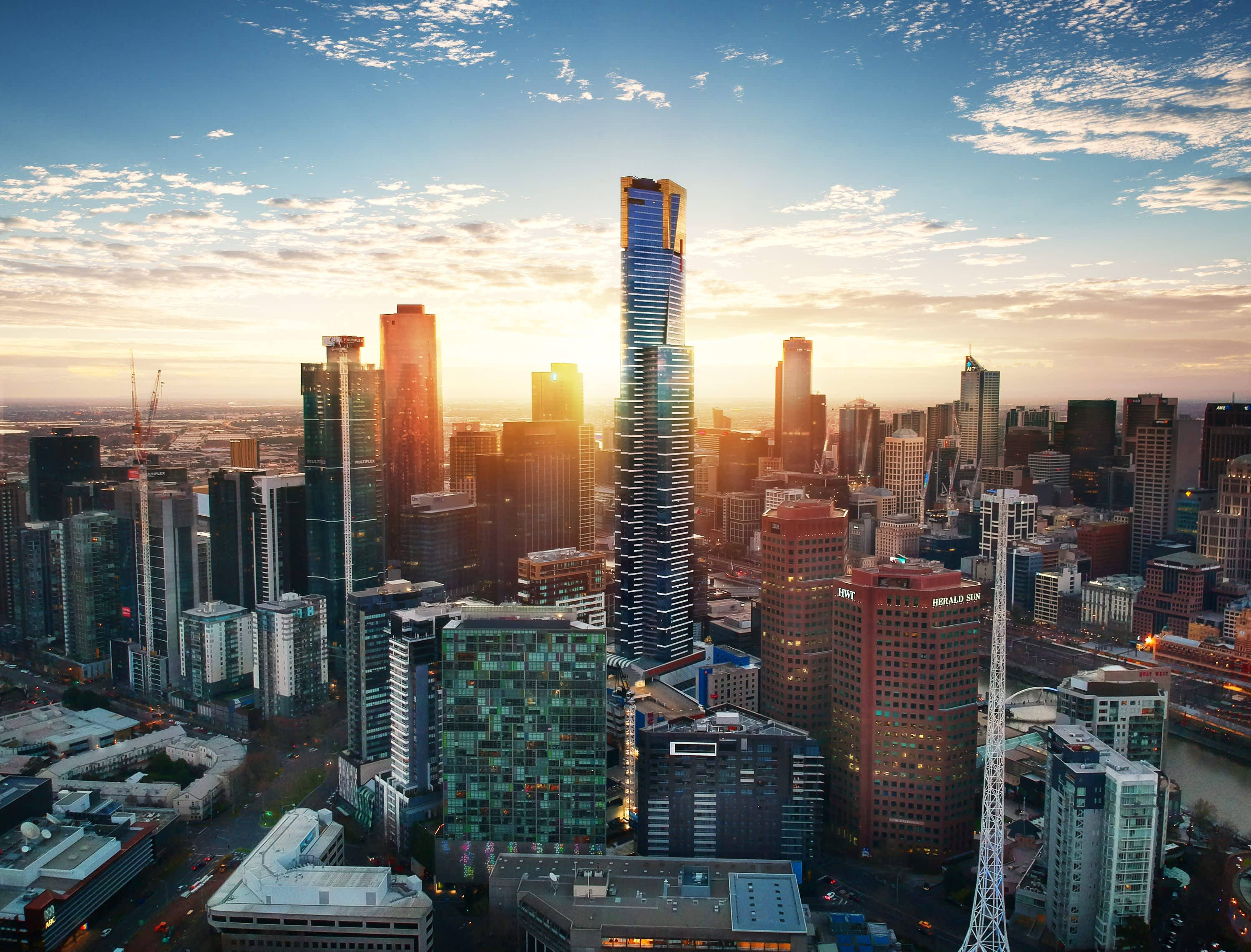 Melbourne 桌布 壁紙 — 現代城市天際線的鳥瞰圖，夕陽西下，金色光芒照亮摩天大樓和散落的雲朵。