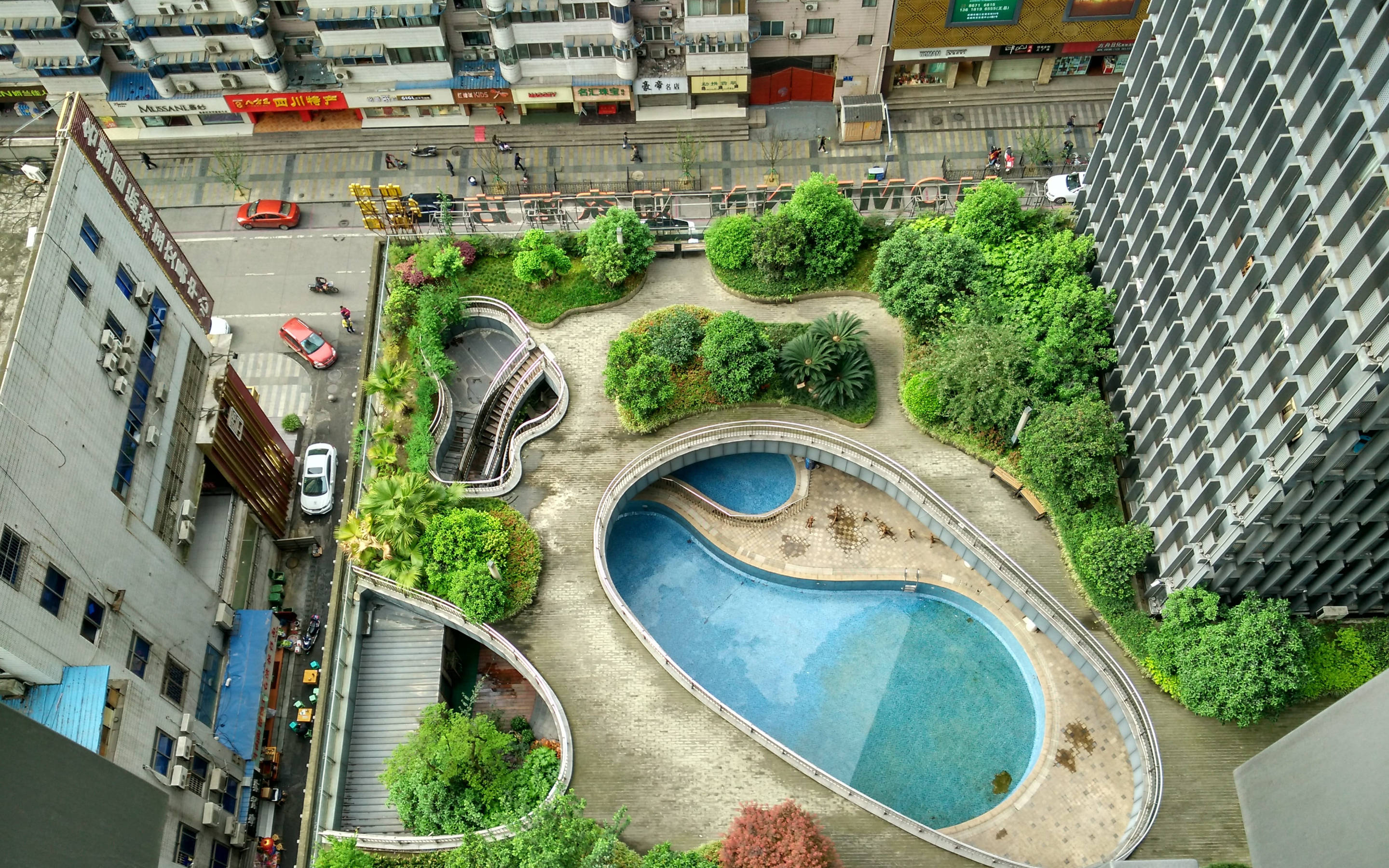 Chengdu 桌布 壁紙 — 現代城市庭院的鳥瞰圖，有一個蜿蜒的游泳池，周圍環繞著茂密的綠植，兩旁是高樓大廈，下方可見街道活動。