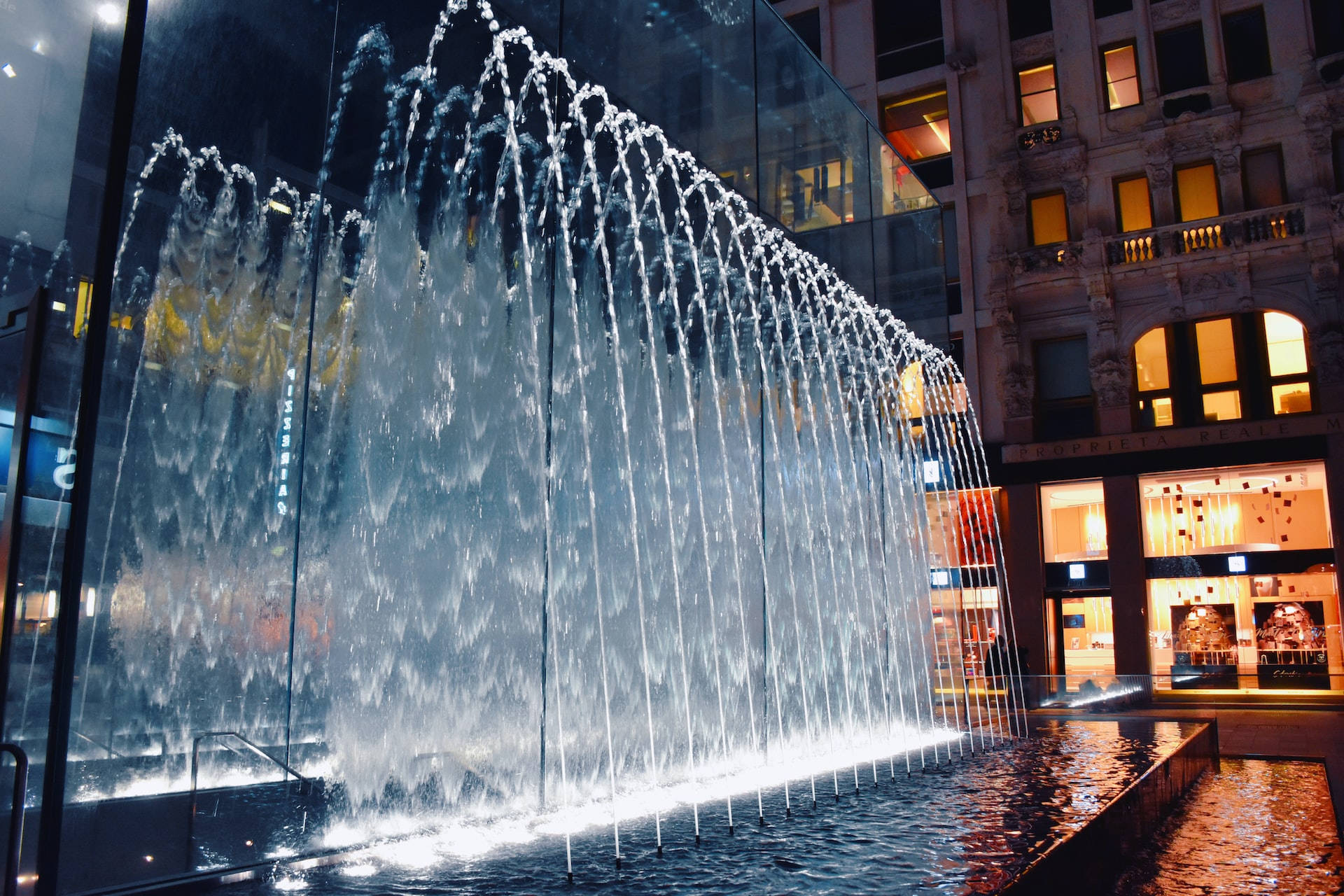 Moving Water 桌布 壁紙 — 現代城市廣場中夜間照亮的層疊瀑布噴泉，背後是發光的建築窗戶。
