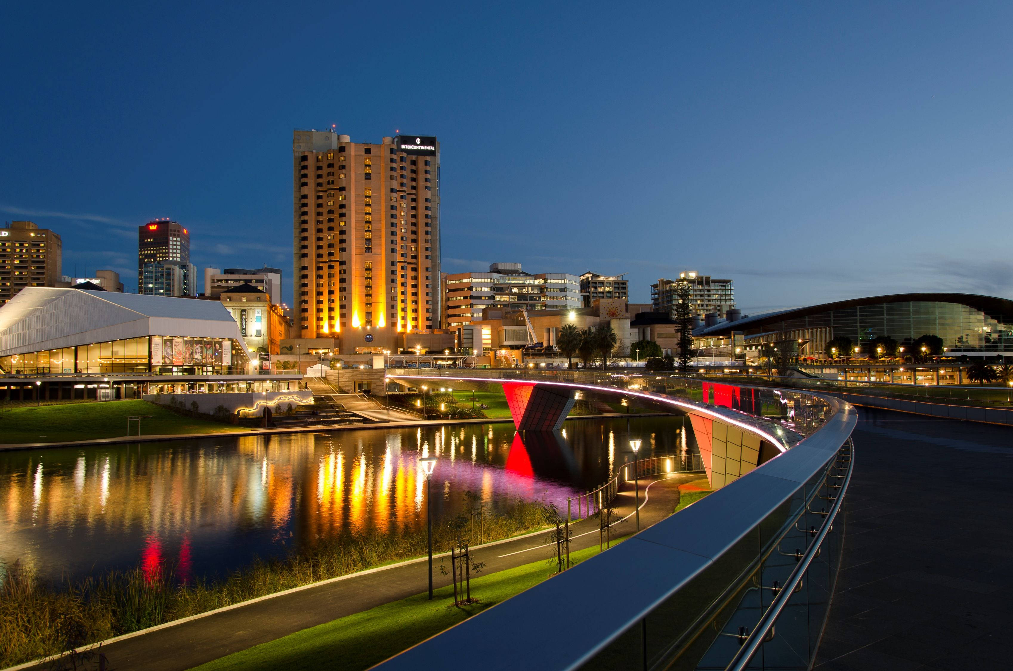 Adelaide 桌布 壁紙 — 現代城市景觀於黃昏時分，建築物燈火通明，一座發光的紅色橋樑倒映在平靜的水面上。