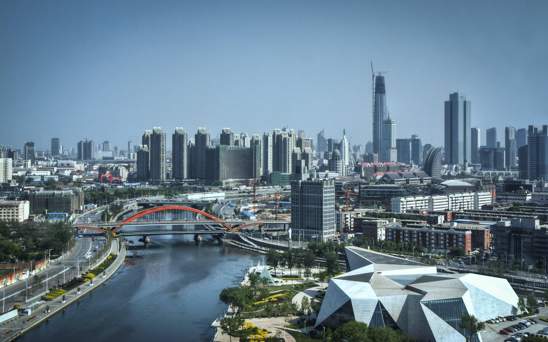 Tianjin 桌布 壁紙 — 現代城市景觀的全景視圖，河流蜿蜒穿過，一座引人注目的紅色拱橋在清澈的藍天下連接城市各區。