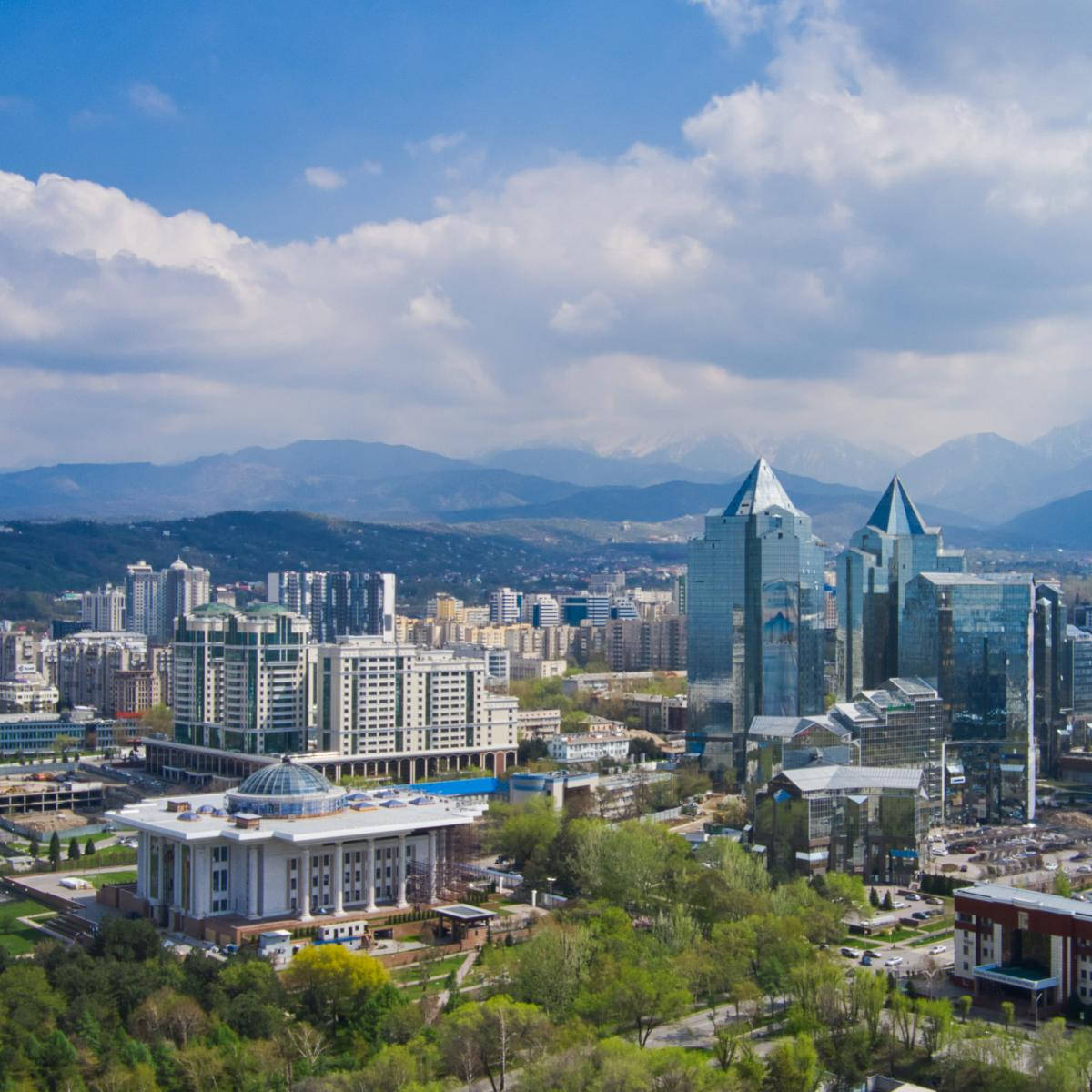 Kazakhstan 桌布 壁紙 — 現代城市景觀的鳥瞰圖，玻璃摩天大樓和綠色公園在明亮的藍天下，有蓬鬆的雲朵和遠處的雪山。