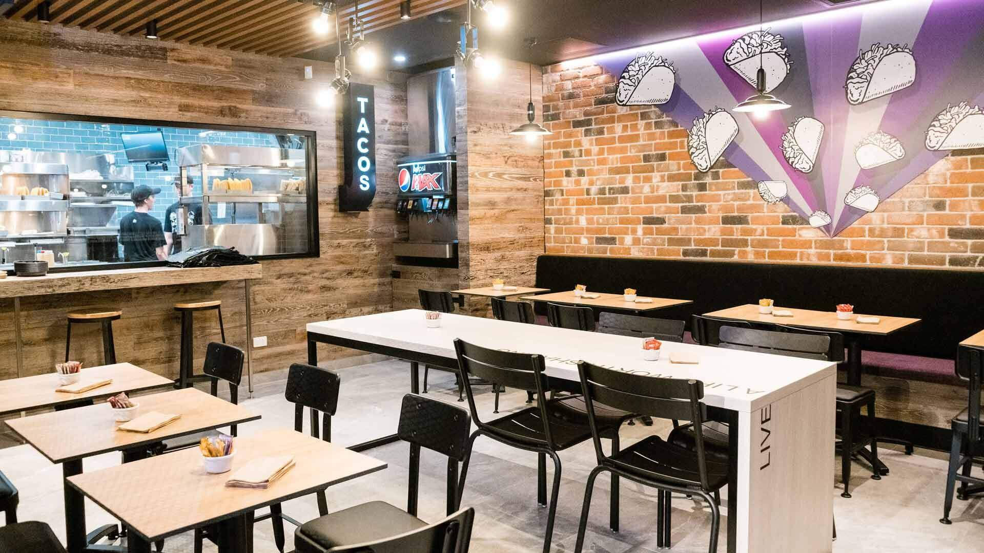 塔可鐘 桌布 壁紙 — 現代墨西哥捲餅餐廳內部，設有磚牆、壁畫、發光燈飾和溫暖吊燈下的木桌。