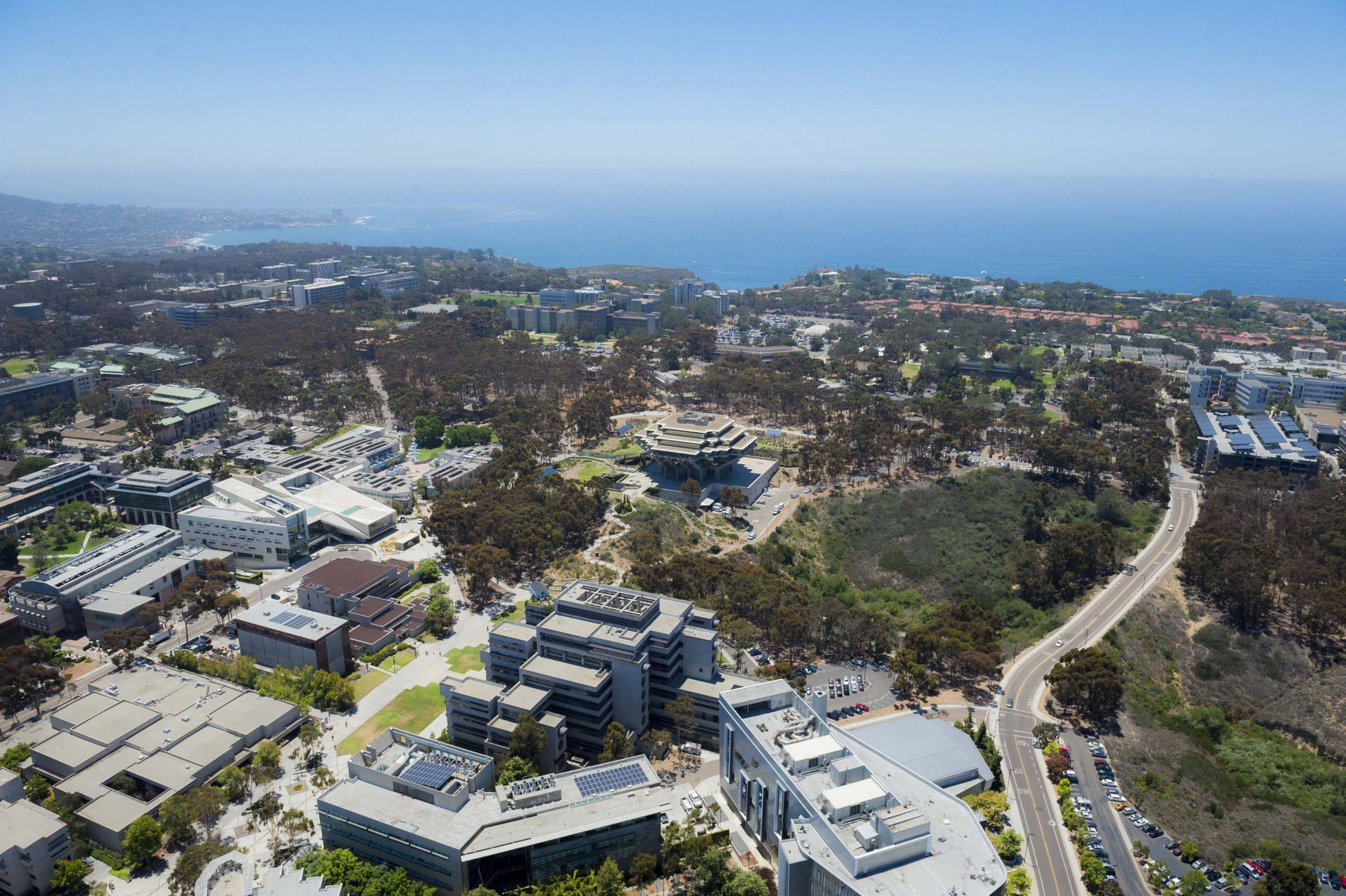 California 桌布 壁紙 — 現代大學校園的鳥瞰圖，坐落在樹木之間，背景是海洋海岸線，天空湛藍清澈。
