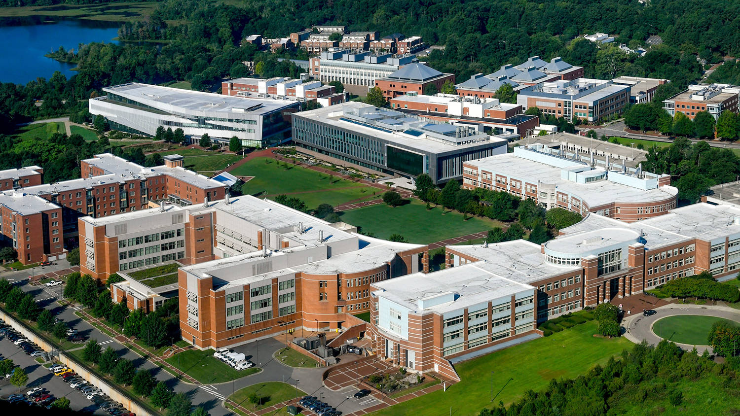 North Carolina 桌布 壁紙 — 現代大學校園的鳥瞰圖，磚和玻璃建築被綠色草坪和樹木環繞，靠近湖泊。