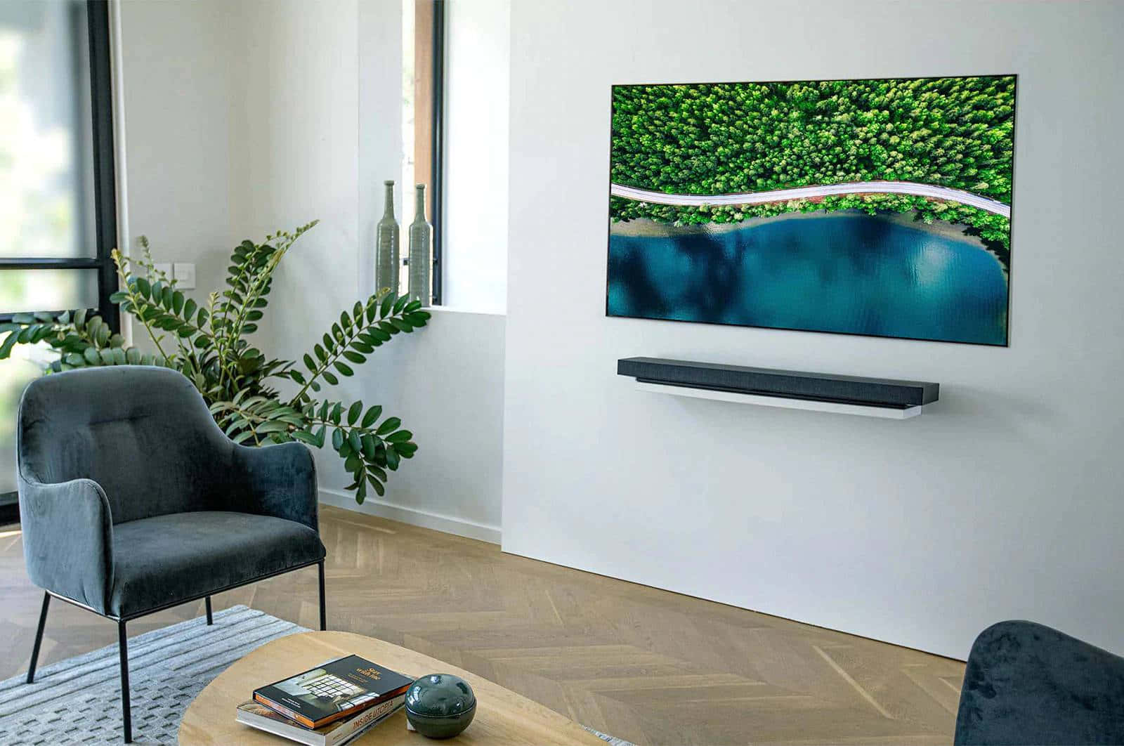 tv 桌布 壁紙 — 現代客廳配灰色天鵝絨椅和人字紋木地板，壁掛電視顯示蜿蜒道路穿過茂密綠色森林和碧綠湖泊的鳥瞰圖。