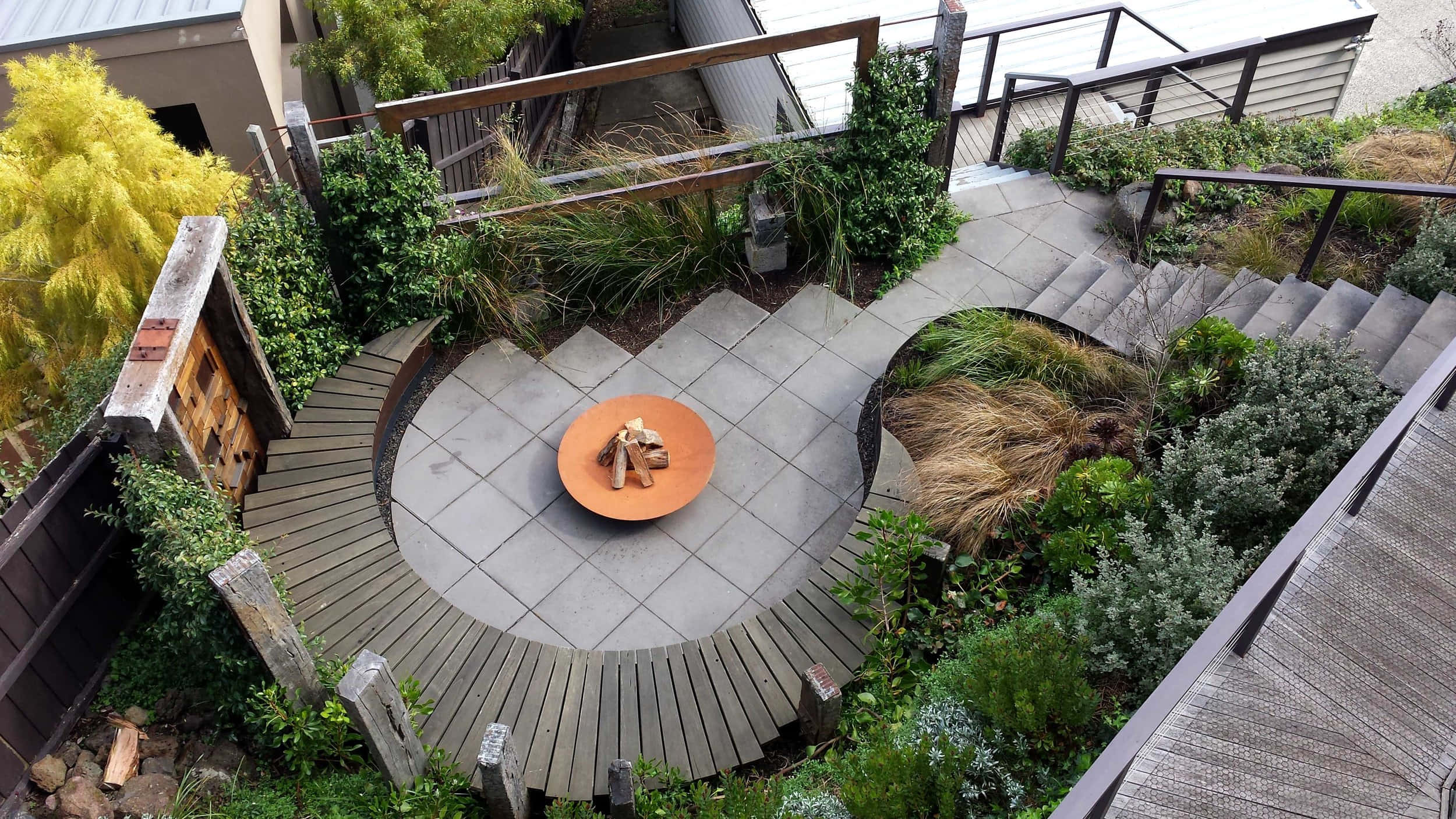 Garden 桌布 壁紙 — 現代屋頂花園的鳥瞰圖，設有弧形木甲板、石磚和中央橙色火坑，周圍環繞著茂密的綠色植物。