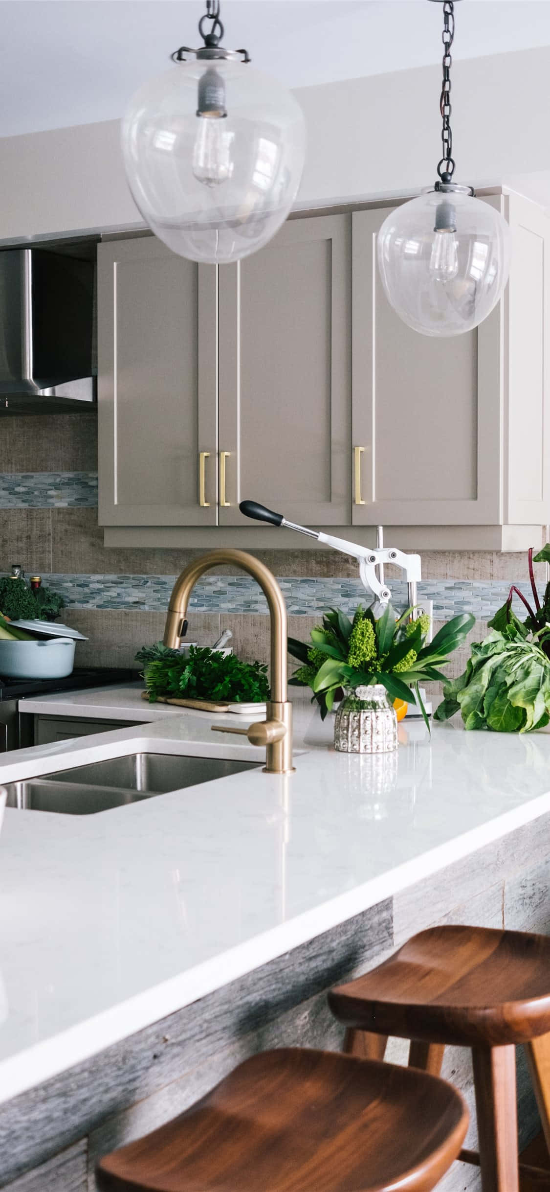 Kitchen Island 桌布 壁紙 — 現代廚房，配有白色石英檯面、黃銅水龍頭、玻璃吊燈和木凳，周圍環繞著茂盛的綠色植物。