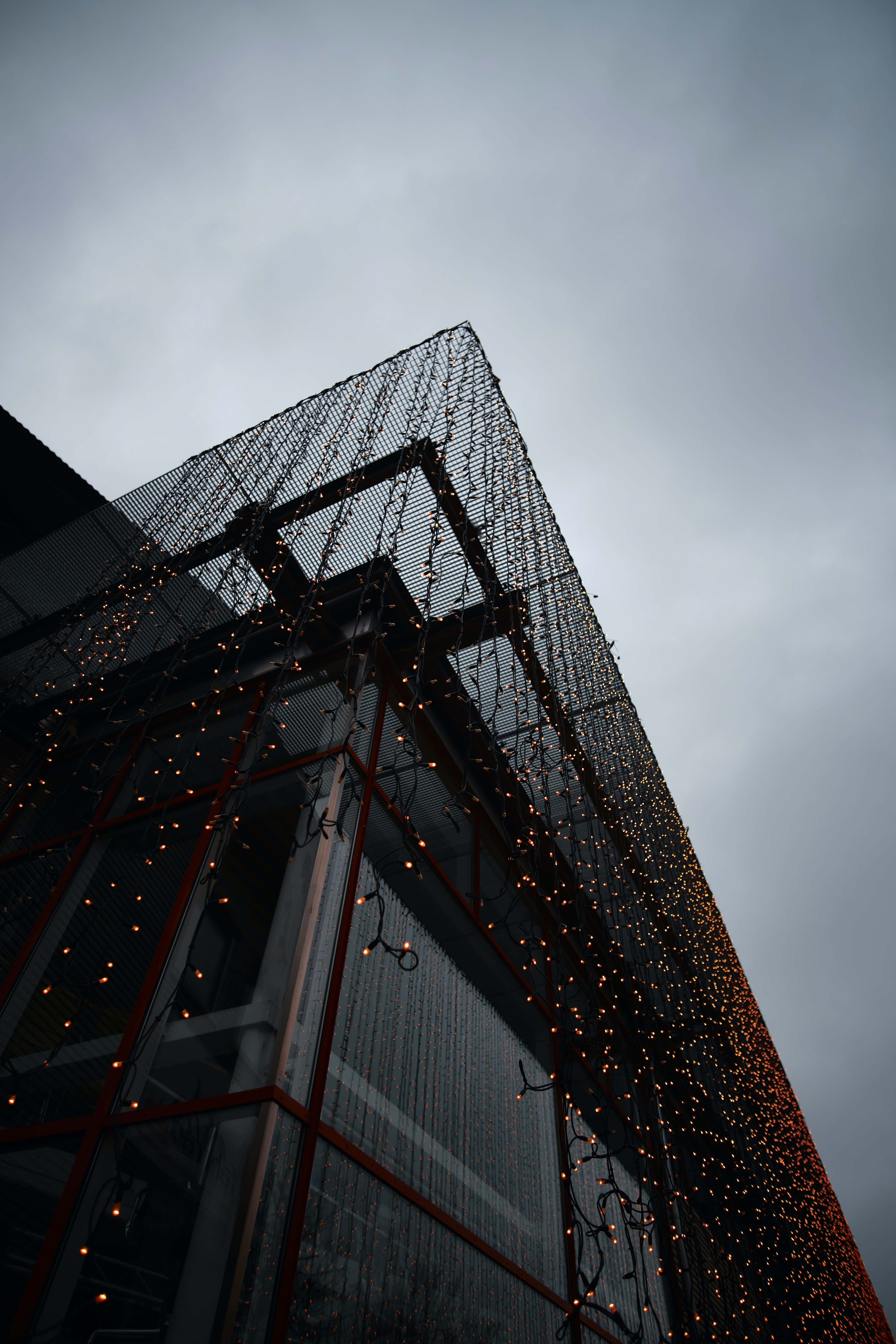 Christmas Light 桌布 壁紙 — 現代建築的玻璃外牆點綴著溫暖的串燈，背景是陰沉的多雲天空。