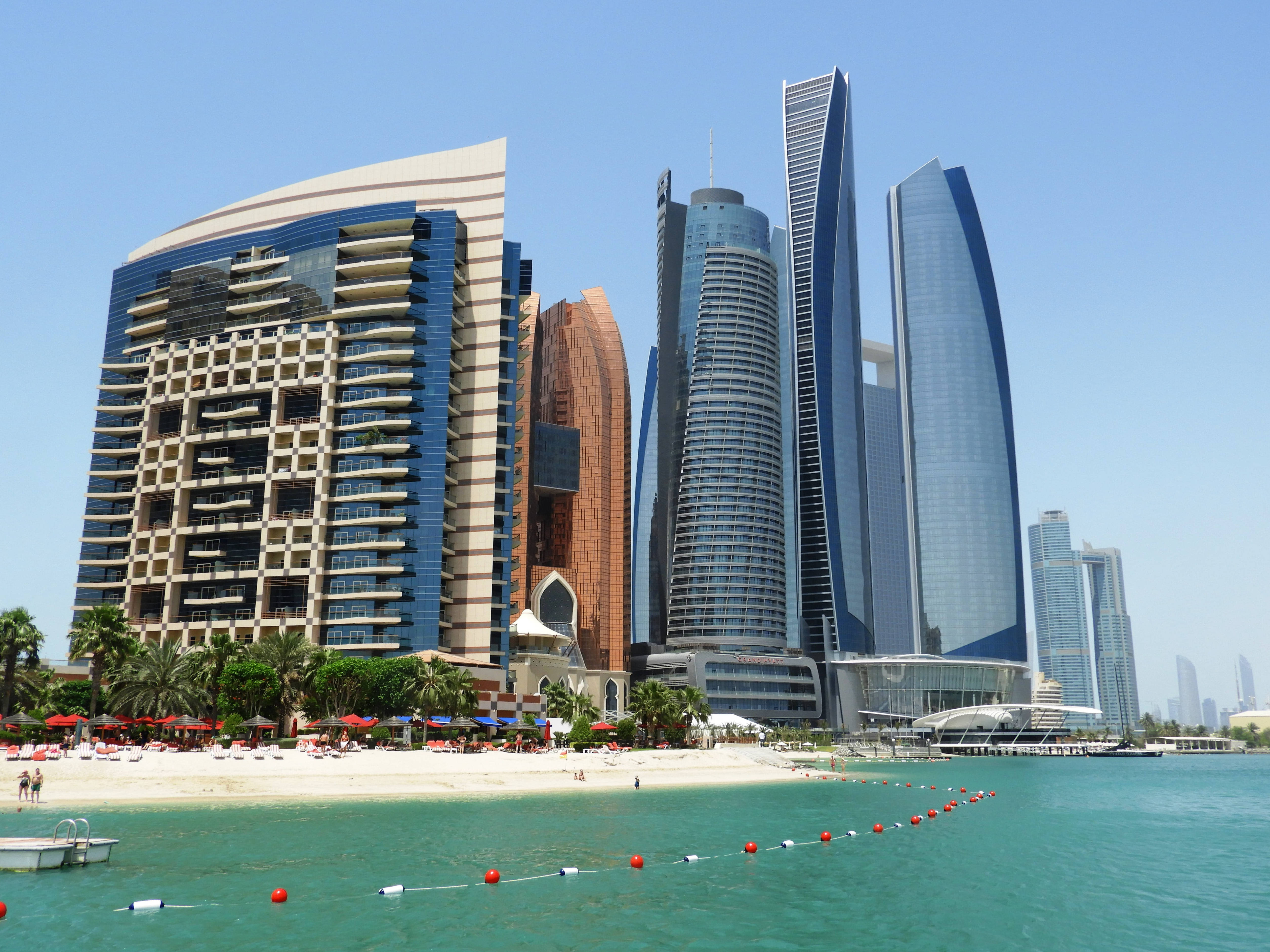 Arab 桌布 壁紙 — 現代摩天大樓以玻璃和銅質外牆矗立在沙灘後方，沙灘上有碧綠海水和紅色遮陽傘，天空清澈湛藍。