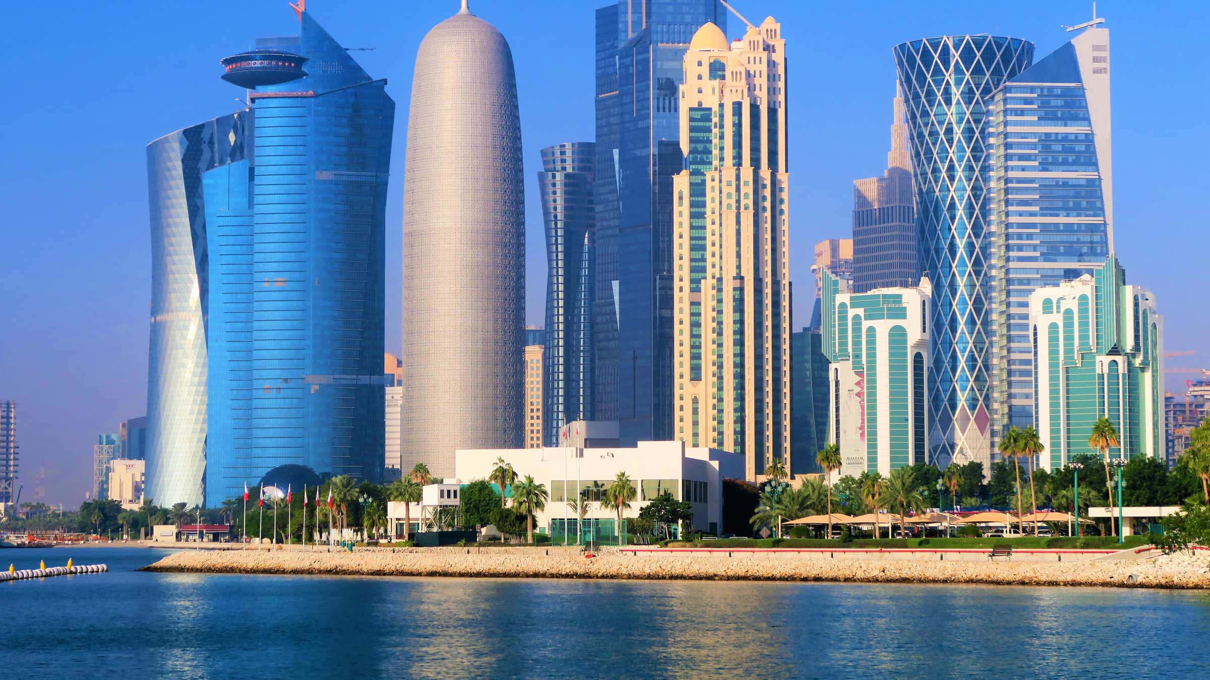 Qatar 桌布 壁紙 — 現代摩天大樓以藍色和銀色玻璃幕牆，在晴朗藍天下，聳立於棕櫚樹成蔭的寧靜海濱。