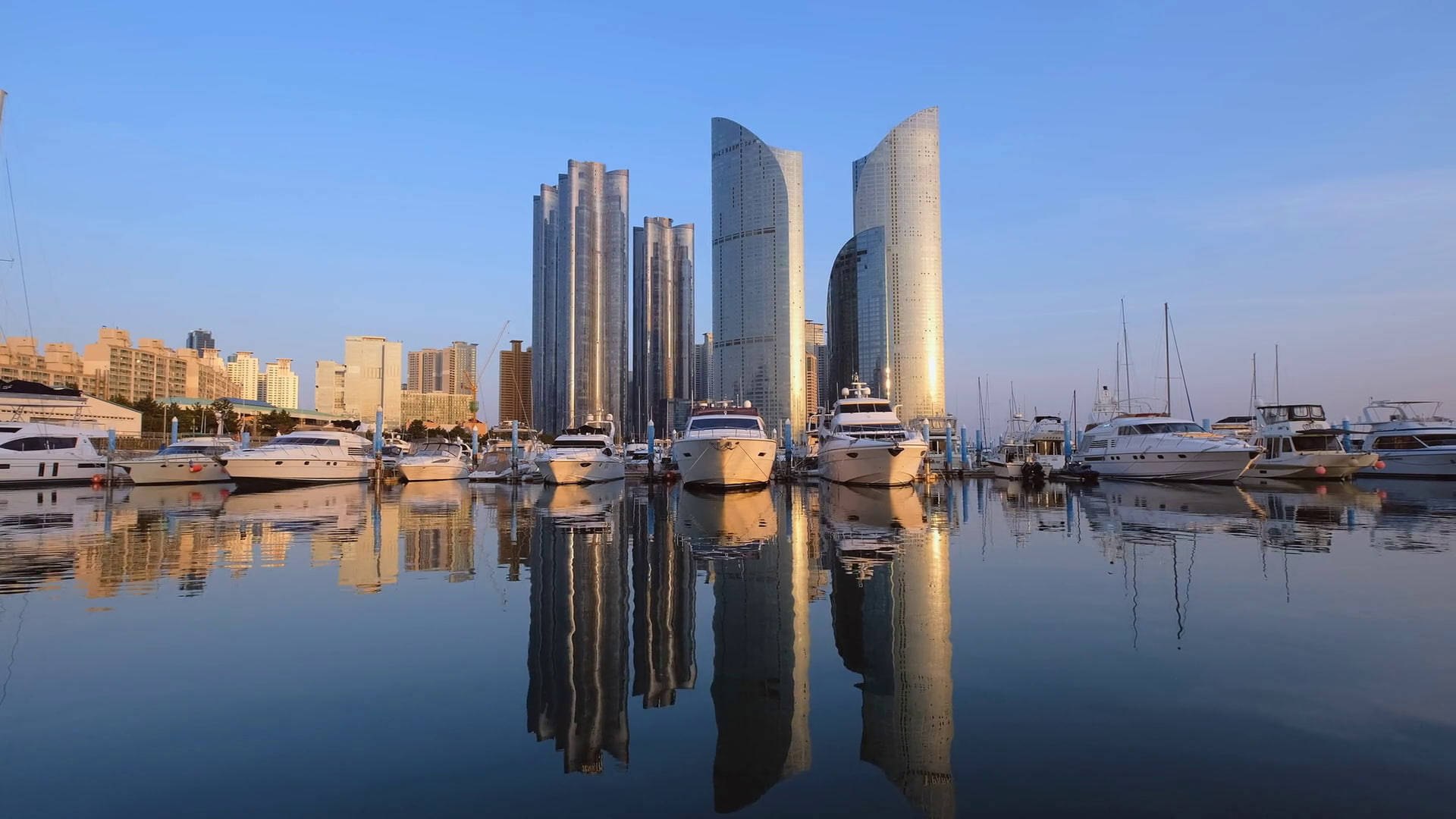Busan 桌布 壁紙 — 現代摩天大樓和豪華遊艇在金色時刻的清澈藍天下，完美倒映在平靜的港灣水面上。