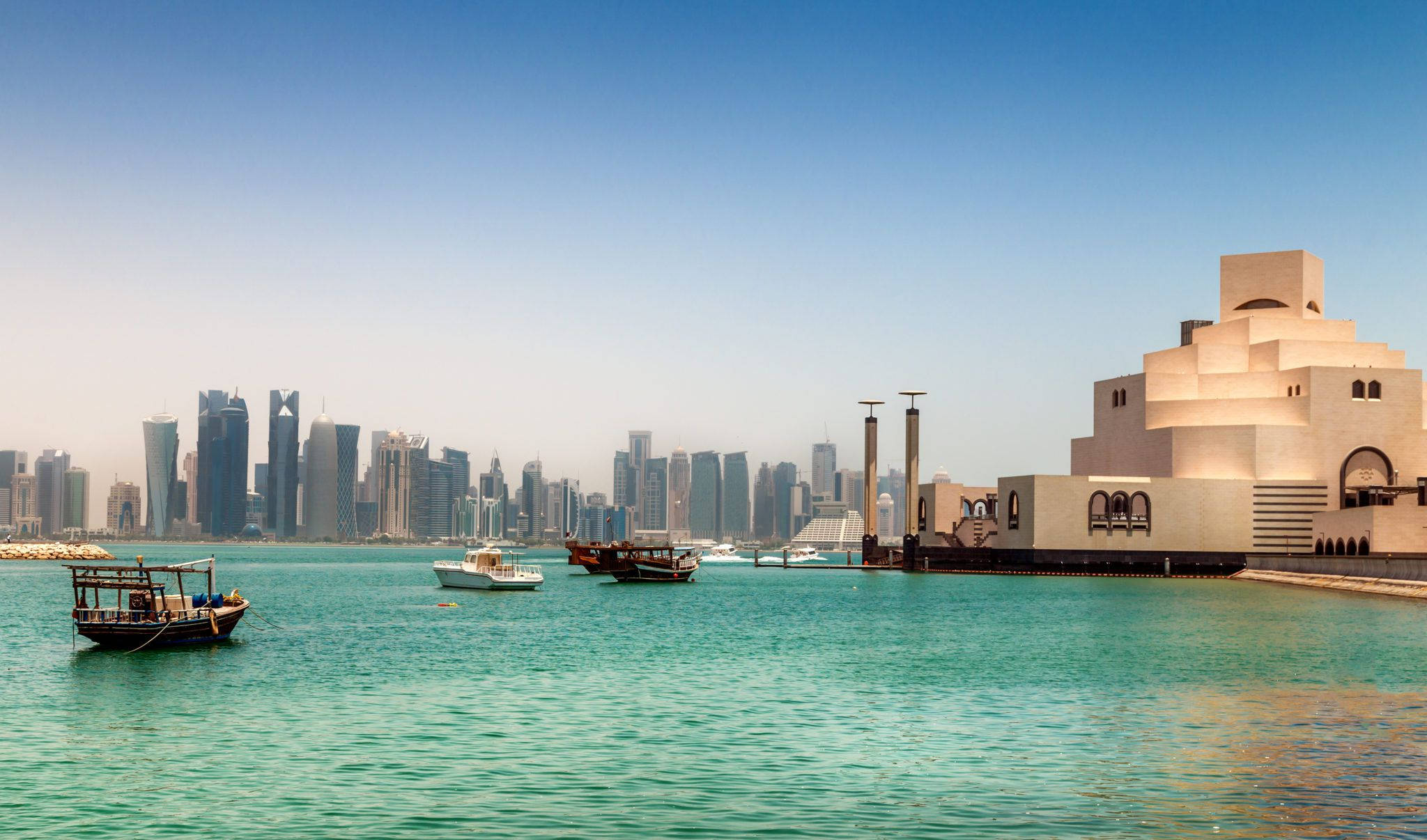 Qatar 桌布 壁紙 — 現代摩天大樓在碧綠海灣後方聳立，傳統木船滑過米色階梯式博物館旁。