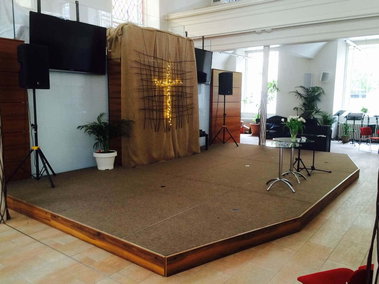 Church Stage 桌布 壁紙 — 現代教堂中一個高起的木製舞台，帶有發光的十字架背景、揚聲器和盆栽植物，在明亮的日光下。