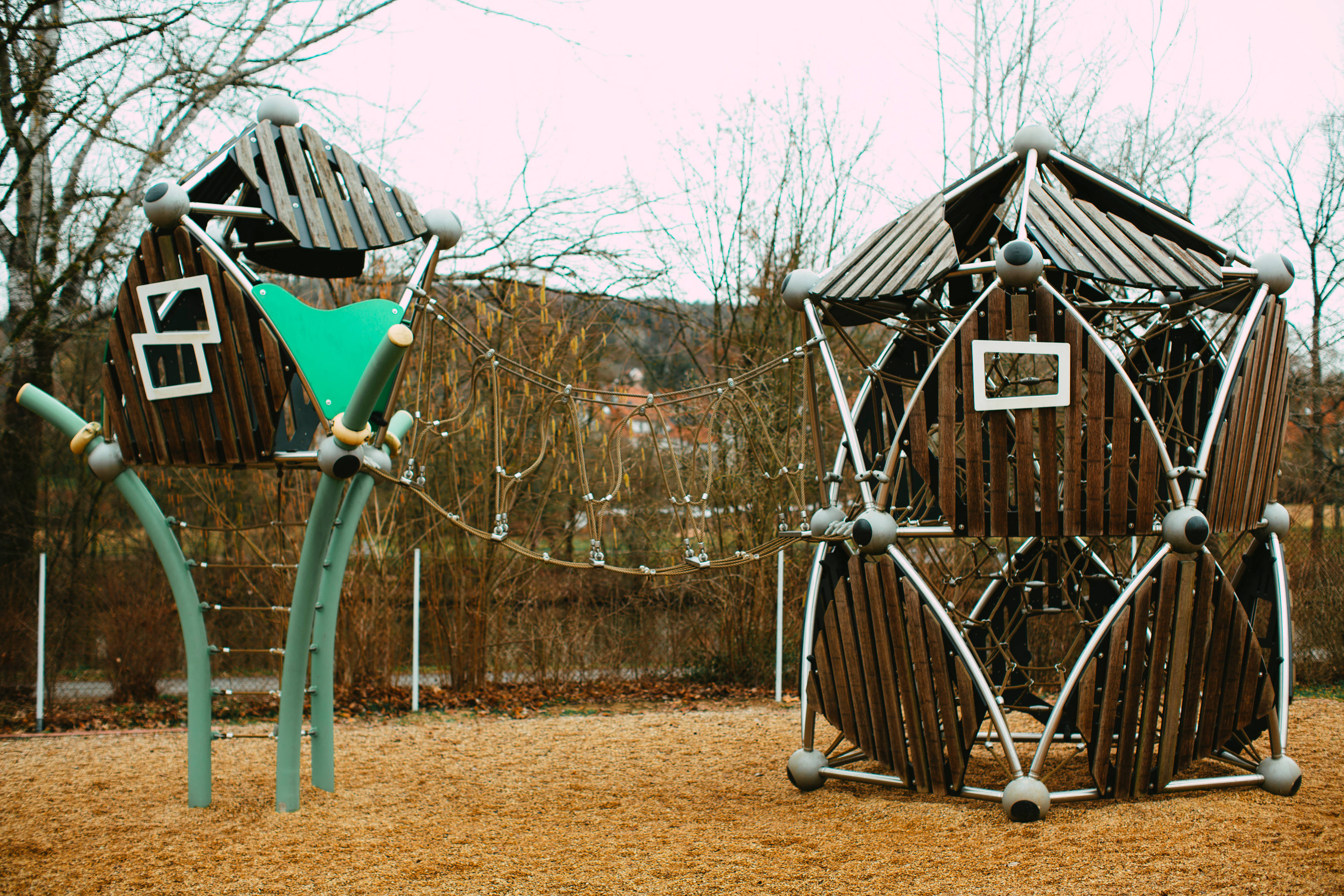Playground 桌布 壁紙 — 現代木製遊樂場結構，配有綠色溜滑梯和繩橋，立於木屑地面上，周圍是光禿禿的樹木。