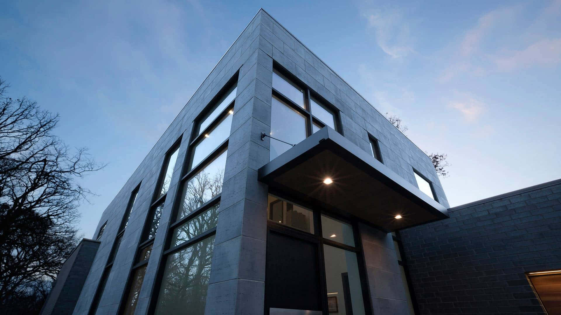 Minimalist Architecture 桌布 壁紙 — 現代棱角建築，灰色石頭和玻璃窗，在暮色天空下，有光禿禿的樹木。