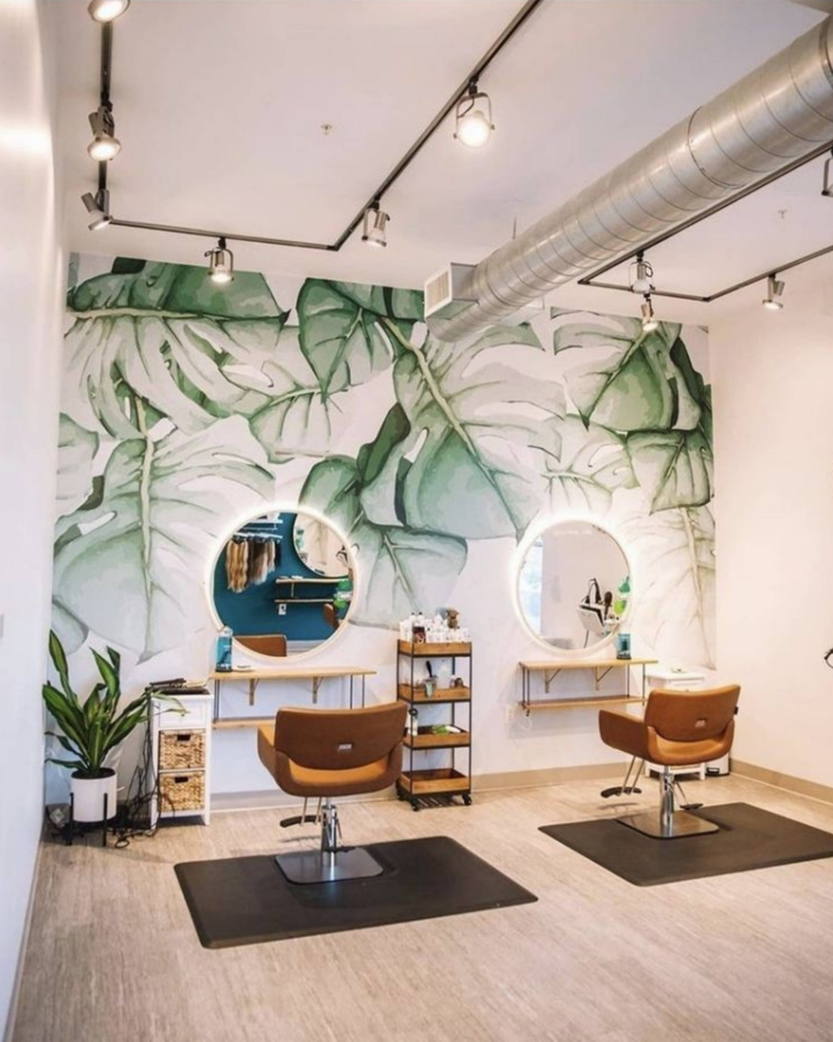 Hair Salon 桌布 壁紙 — 現代沙龍，配有大型熱帶葉子壁紙、兩張棕色椅子、圓形鏡子和明亮燈光下暴露的管道。
