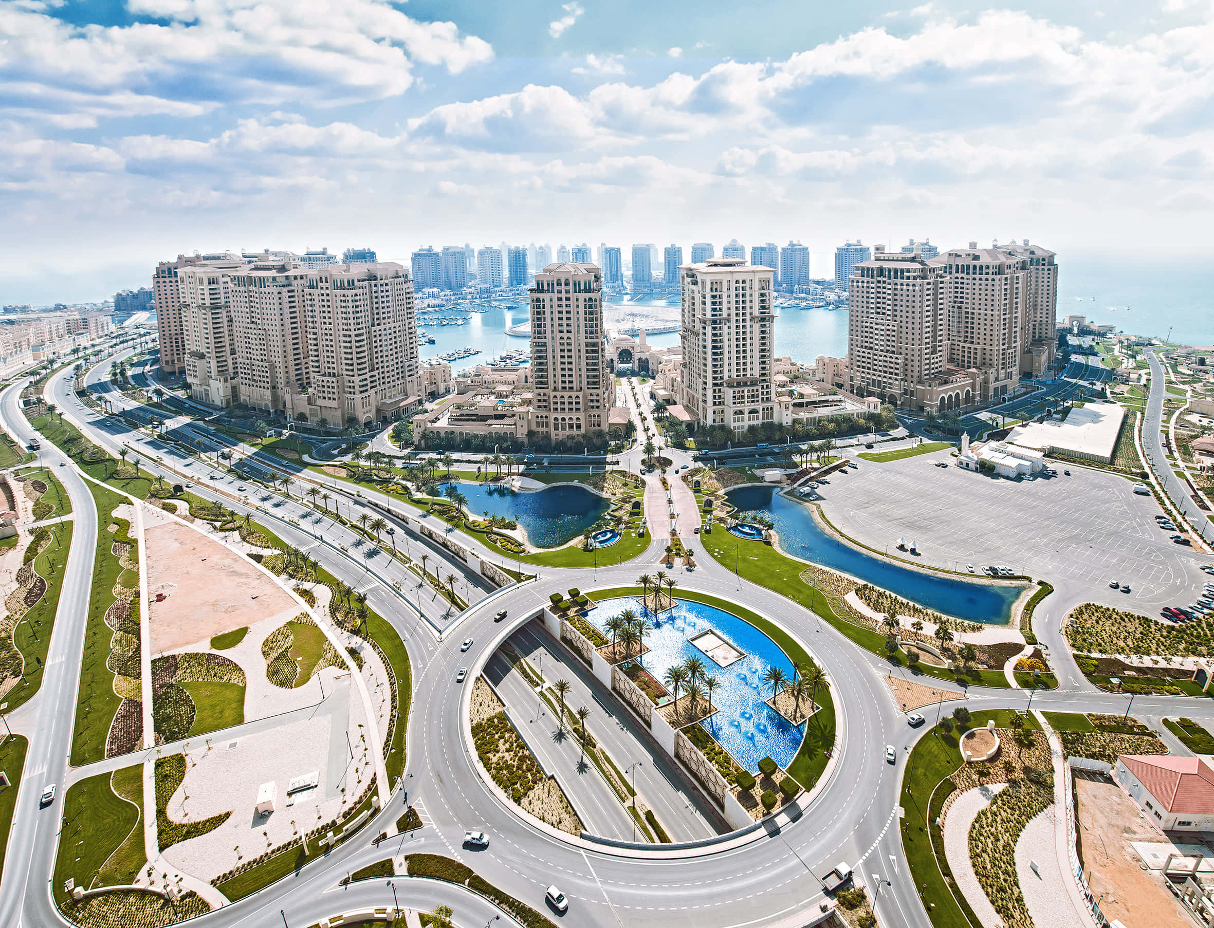 卡達 桌布 壁紙 — 現代沿海城市的鳥瞰圖，彎曲的高速公路蜿蜒穿過園林公園和高層建築，在明亮的藍天和蓬鬆的白雲下。