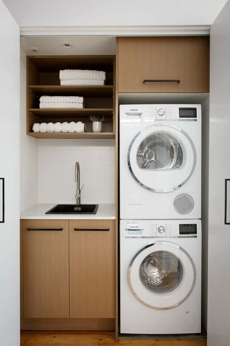 Laundry Room 桌布 壁紙 — 現代洗衣角落，配有堆疊的白色洗衣機和乾衣機，以及存放摺疊毛巾的木製櫥櫃。