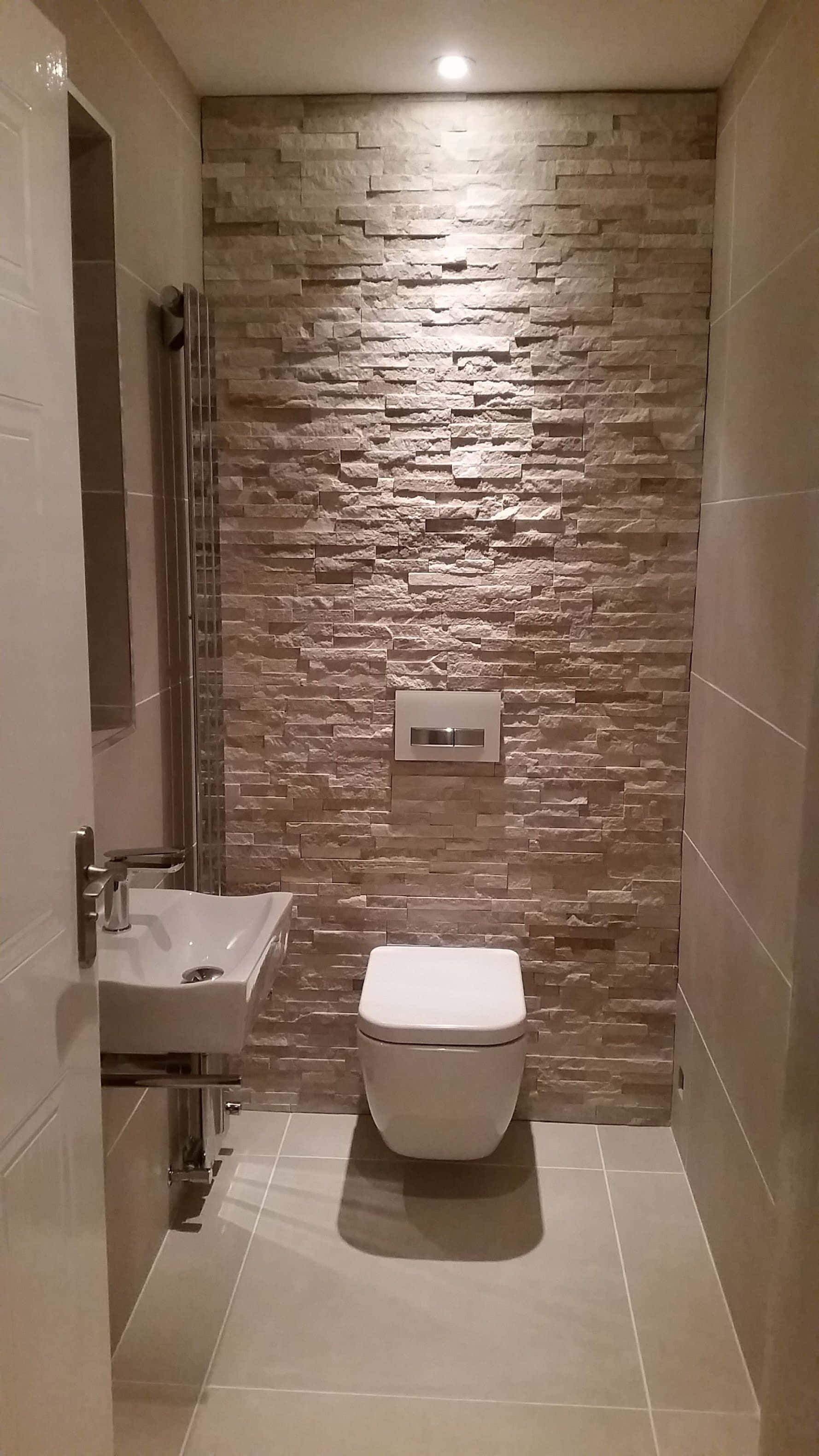 Toilet 桌布 壁紙 — 現代浴室，米色石質特色牆由天花板燈照亮，上方是時尚白色馬桶和洗手台。
