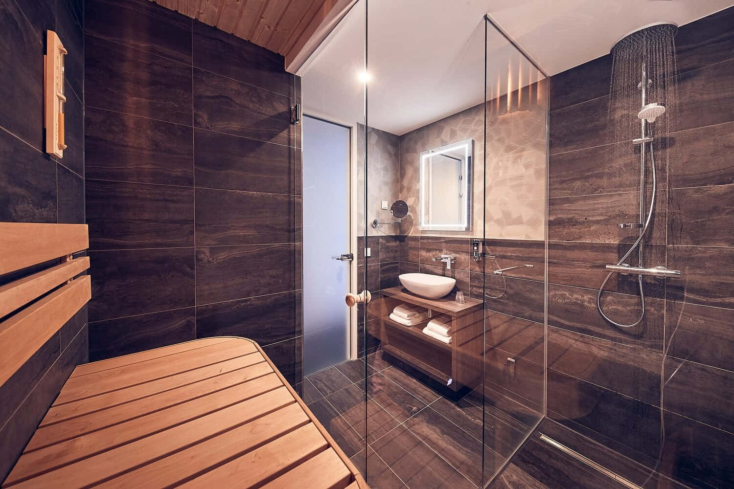 Interior Design 桌布 壁紙 — 現代浴室，配有深色木紋磚、玻璃淋浴間和木製桑拿長凳，在溫暖燈光下。