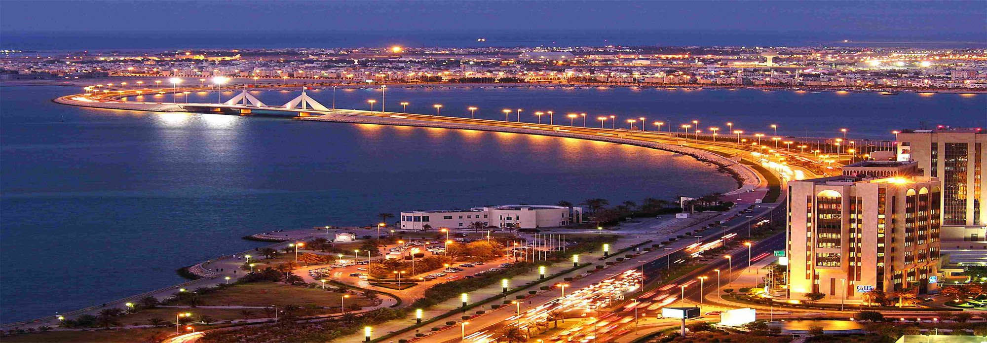 Bahrain 桌布 壁紙 — 現代海岸城市在暮色中的驚人景色，燈光閃爍，倒映在平靜的水面上，還有一座彎曲的照明橋樑。