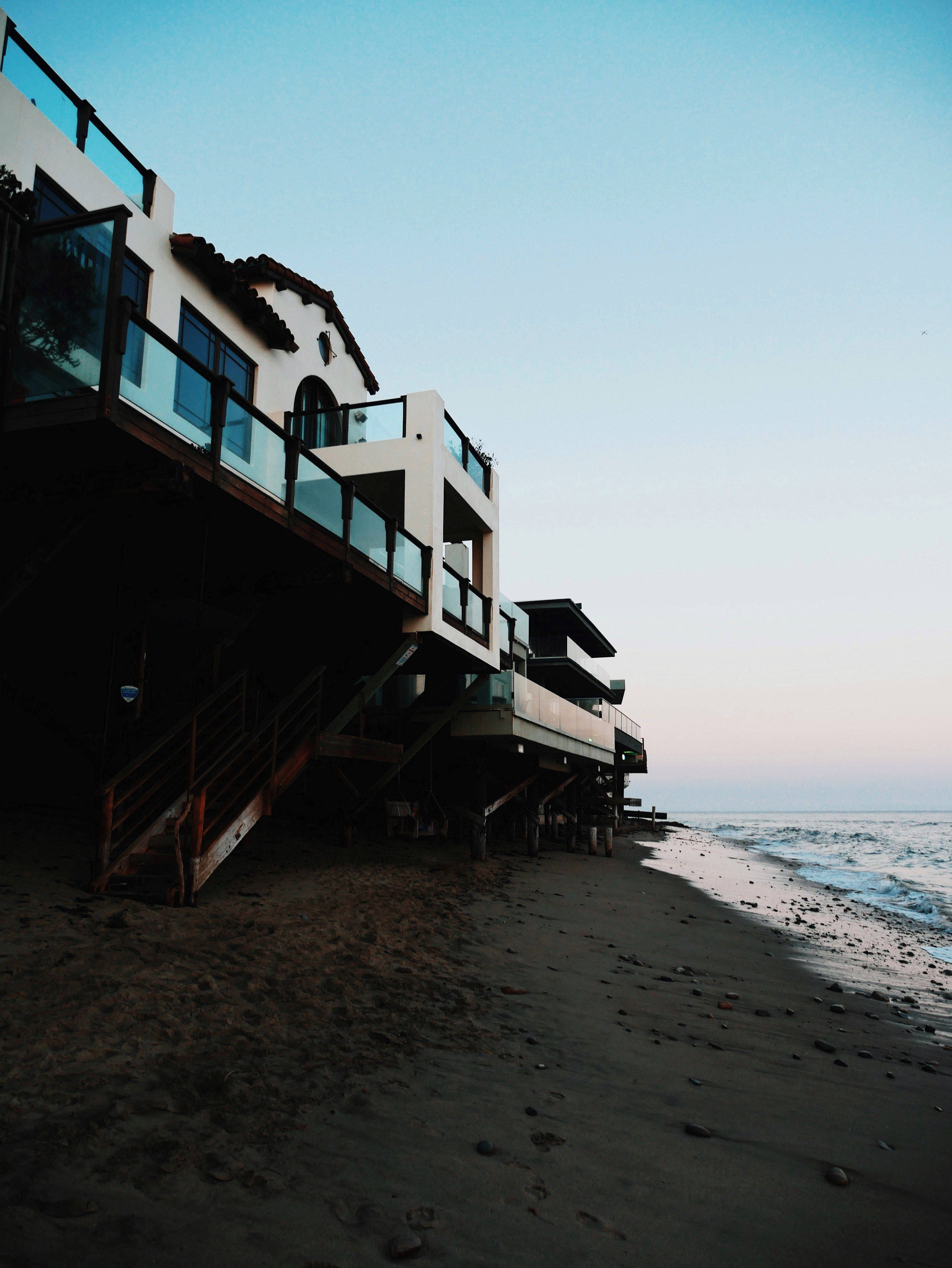 Malibu 桌布 壁紙 — 現代海濱房屋配有玻璃欄杆，矗立在深色沙灘上的高蹺上，溫柔的海浪在黃昏時分湧入。