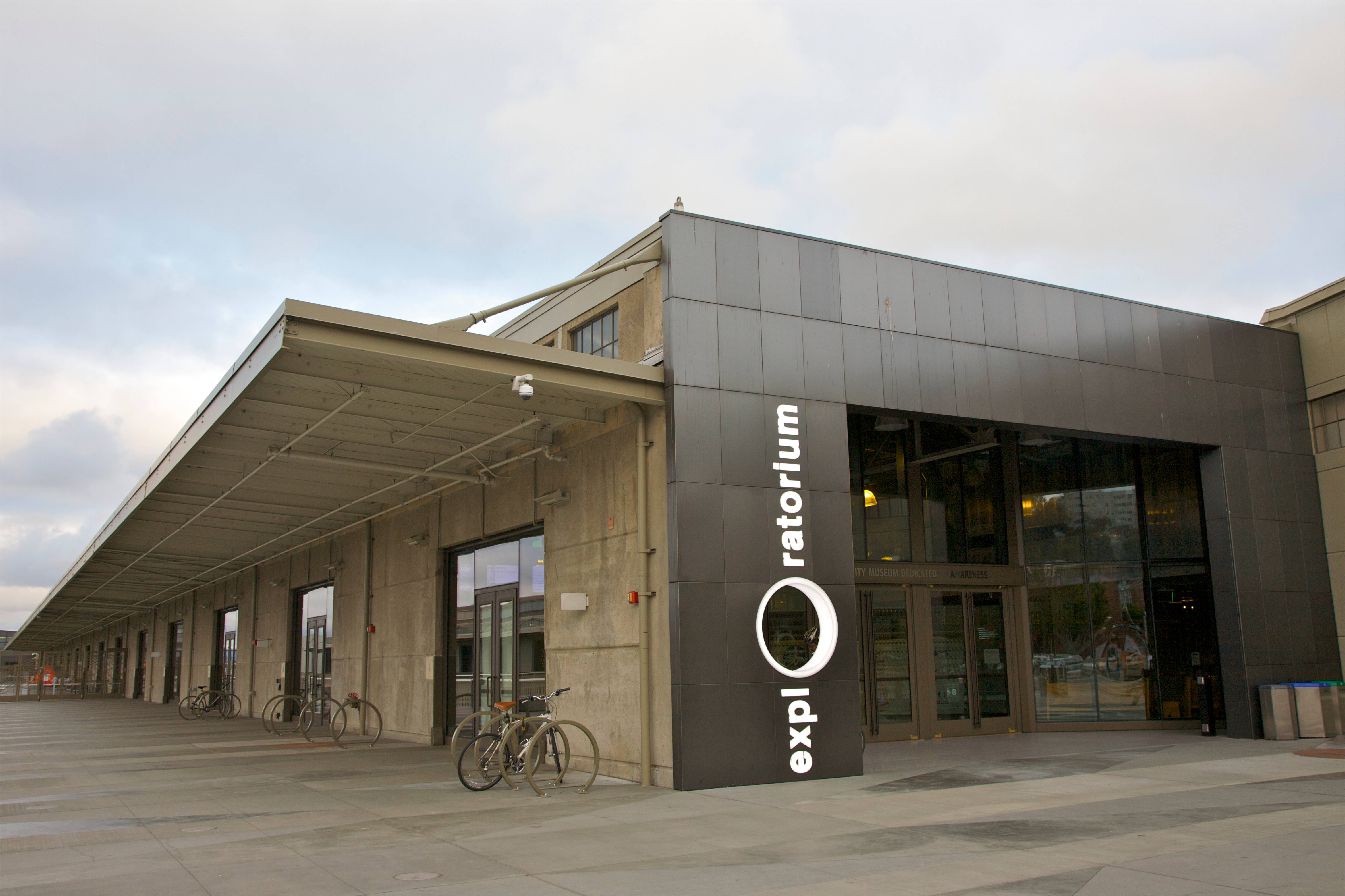 舊金山 桌布 壁紙 — 現代混凝土和玻璃建築，帶有 'exploratorium' 標誌，腳踏車停放在外面，天空多雲。