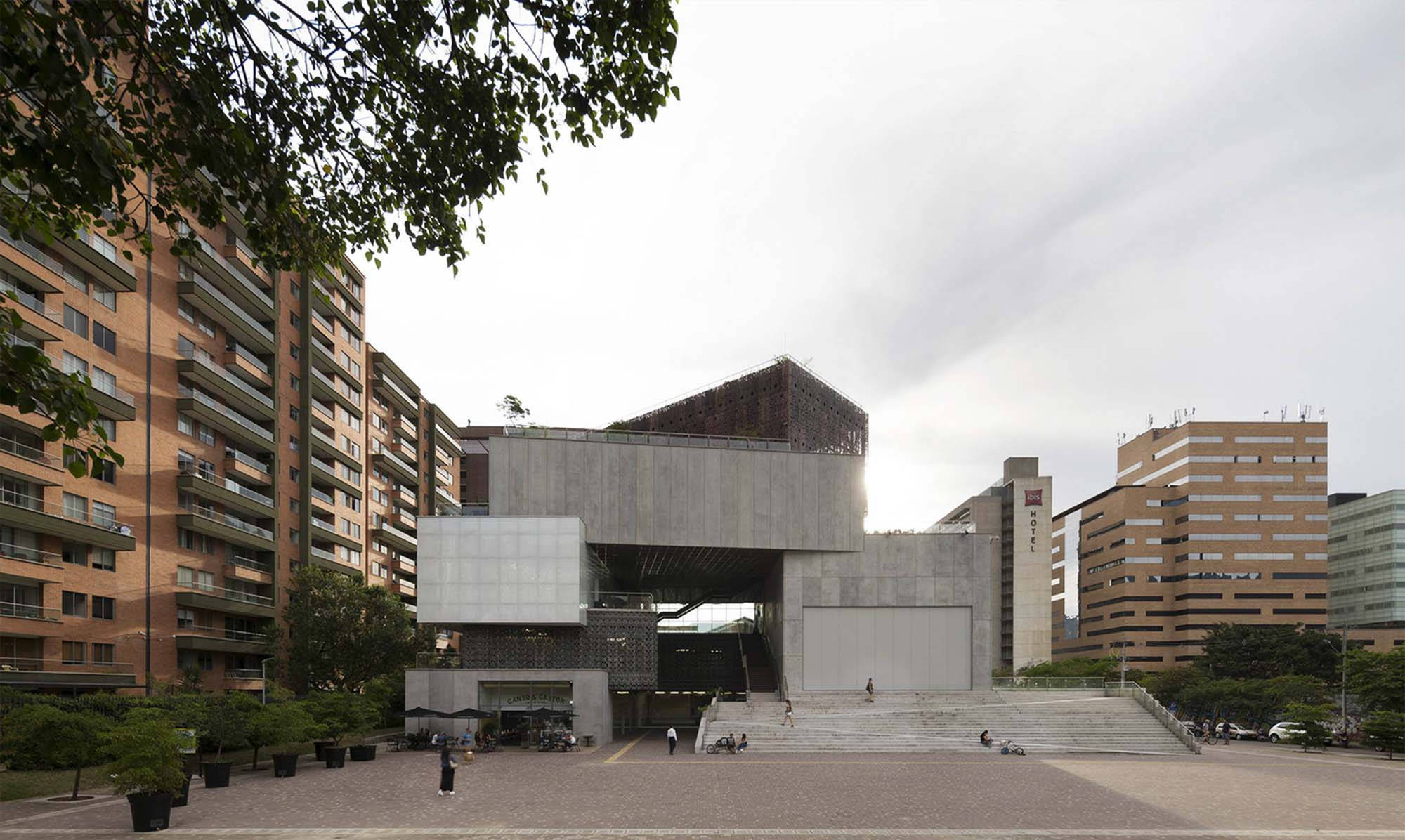 Medellin 桌布 壁紙 — 現代混凝土和玻璃建築結構，幾何形狀，矗立在城市廣場，周圍是住宅建築和綠化，天空多雲。