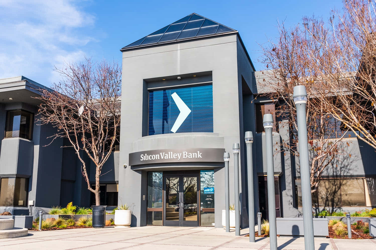 Bank 桌布 壁紙 — 現代灰色銀行大樓，帶有大型箭頭標誌，背景是晴朗藍天和光禿禿的樹木。
