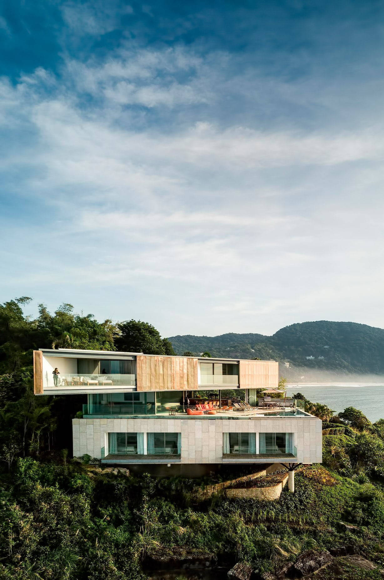 Luxury House 桌布 壁紙 — 現代玻璃和木屋坐落在懸崖上，俯瞰海洋，周圍是鬱鬱蔥蔥的綠色山丘，天空湛藍。