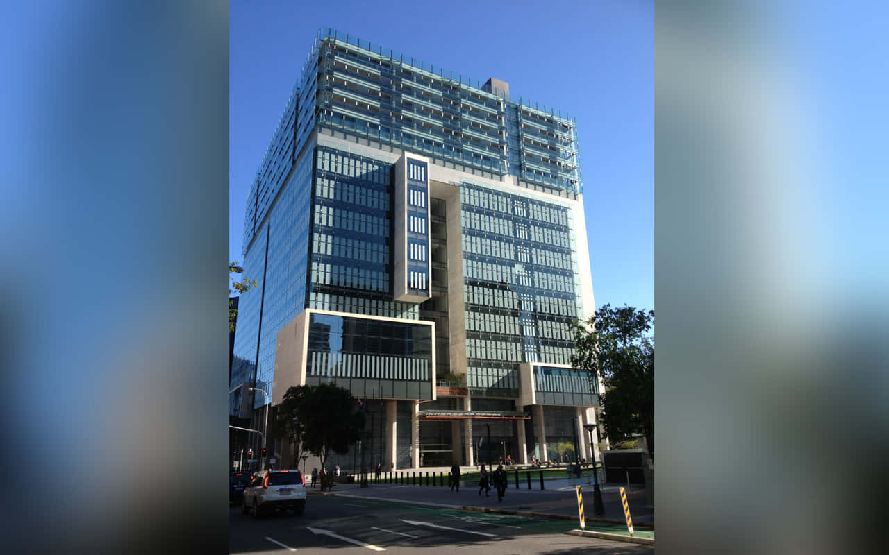 City Building 桌布 壁紙 — 現代玻璃和混凝土摩天大樓在晴朗藍天下聳立於城市街道上，下方有行人和汽車。