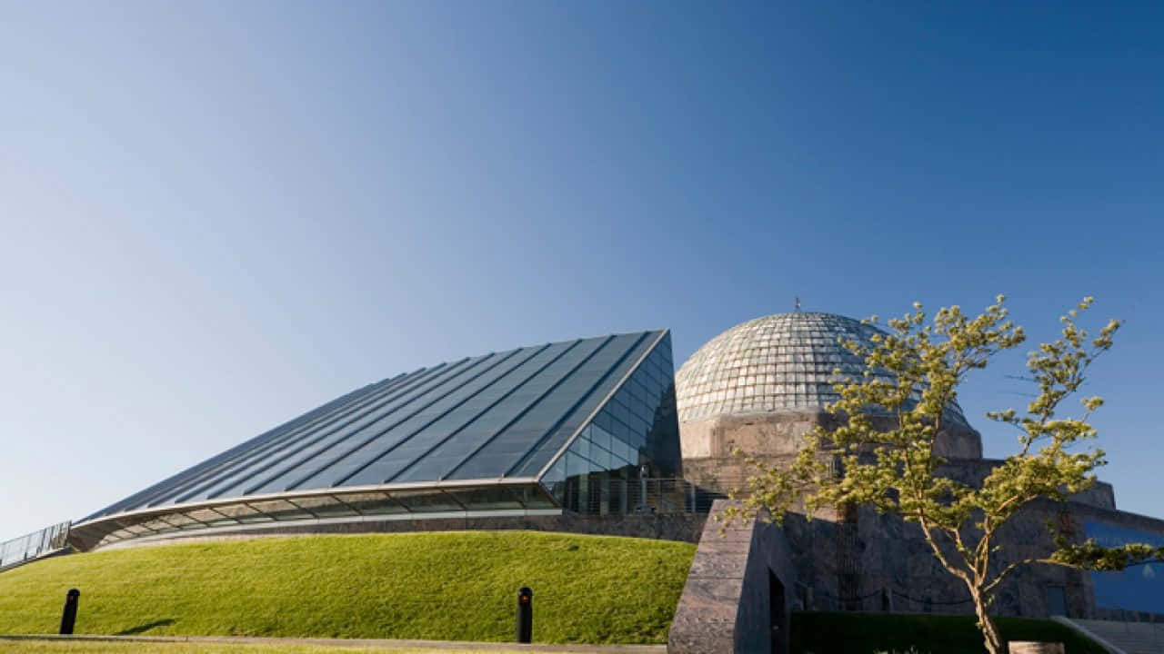 Planetarium 桌布 壁紙 — 現代玻璃和石材建築，帶有圓頂和傾斜屋頂，以清澈的藍天為背景，前景有綠色草地和一棵小樹。