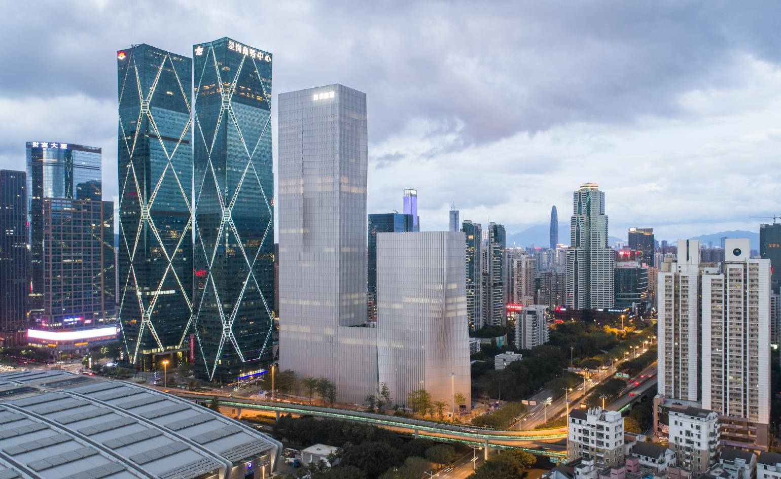 Shenzhen 桌布 壁紙 — 現代玻璃帷幕摩天大樓，帶有幾何圖案，在陰雲密布的暮色天空中聳立於繁華的城市景觀之上。