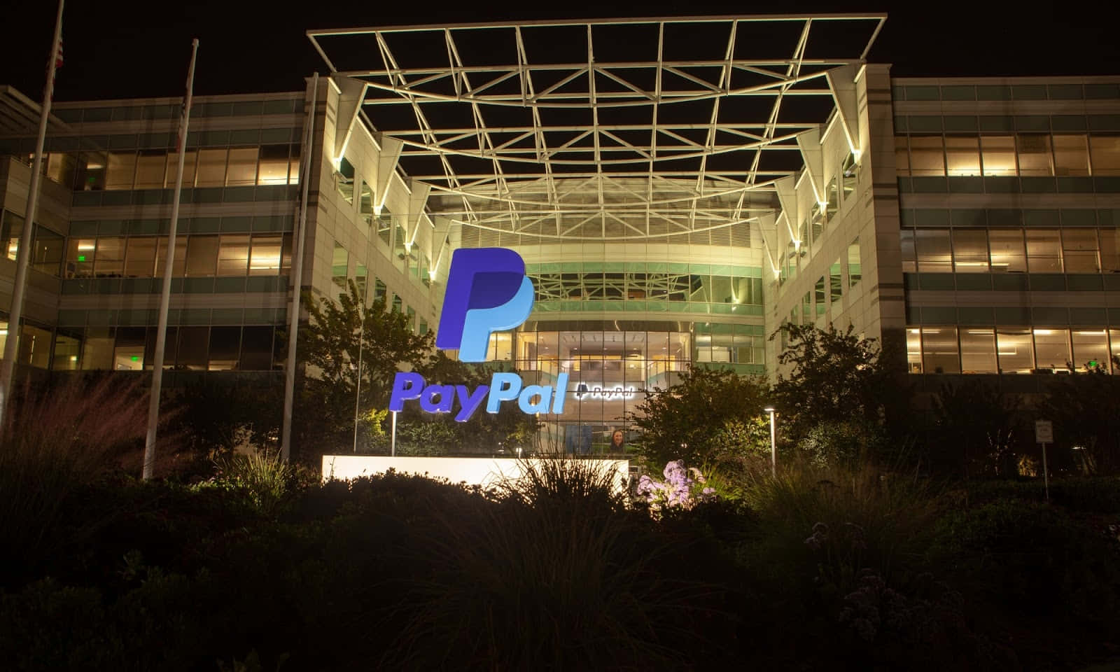 PayPal 桌布 壁紙 — 現代玻璃建築的夜景，帶有發光的PayPal標誌和周圍的樹葉。